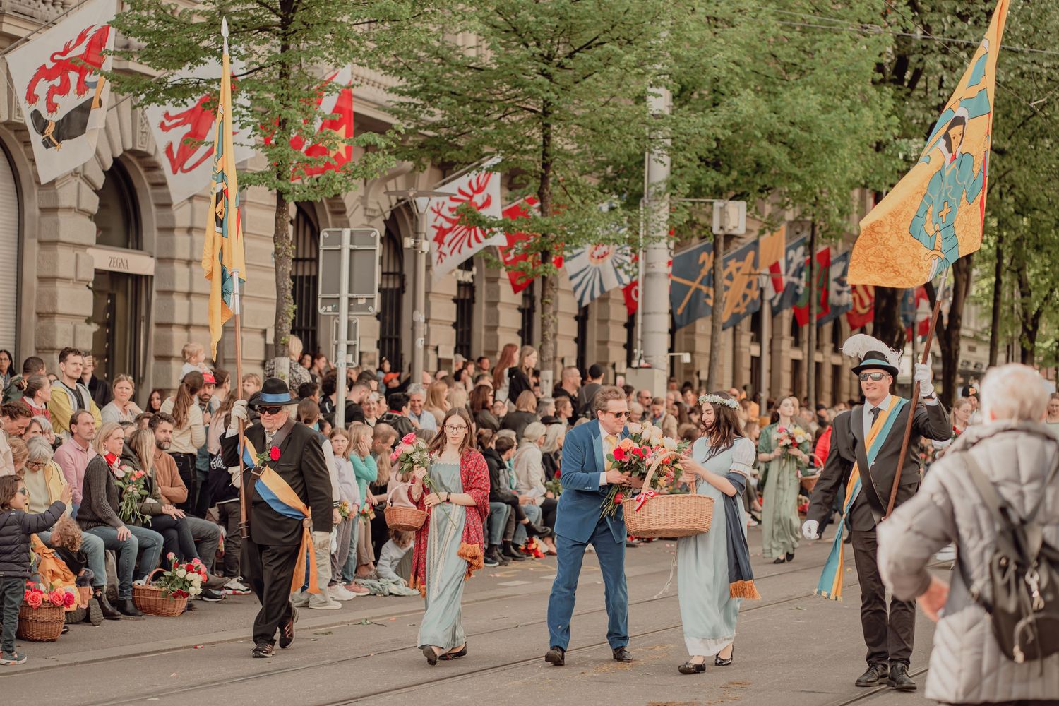 Colorful flag bearers and flower carriers parade past a large crowd at a historic street festival.