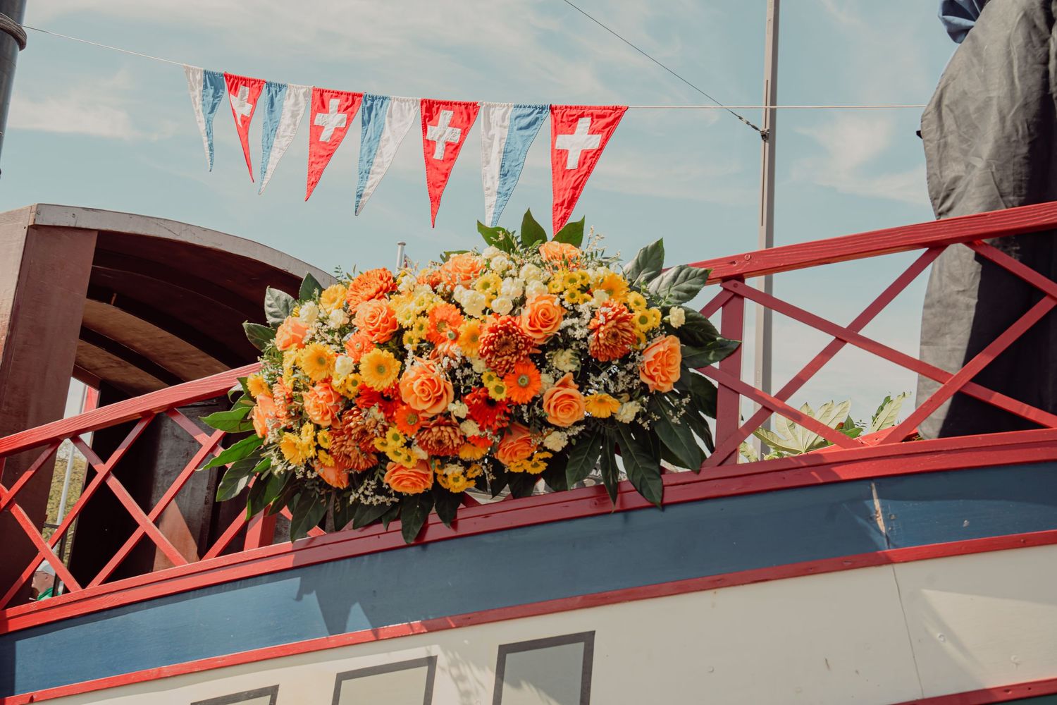 Beautiful orange and yellow floral arrangement displayed on a red railing with Swiss flag pennants overhead.