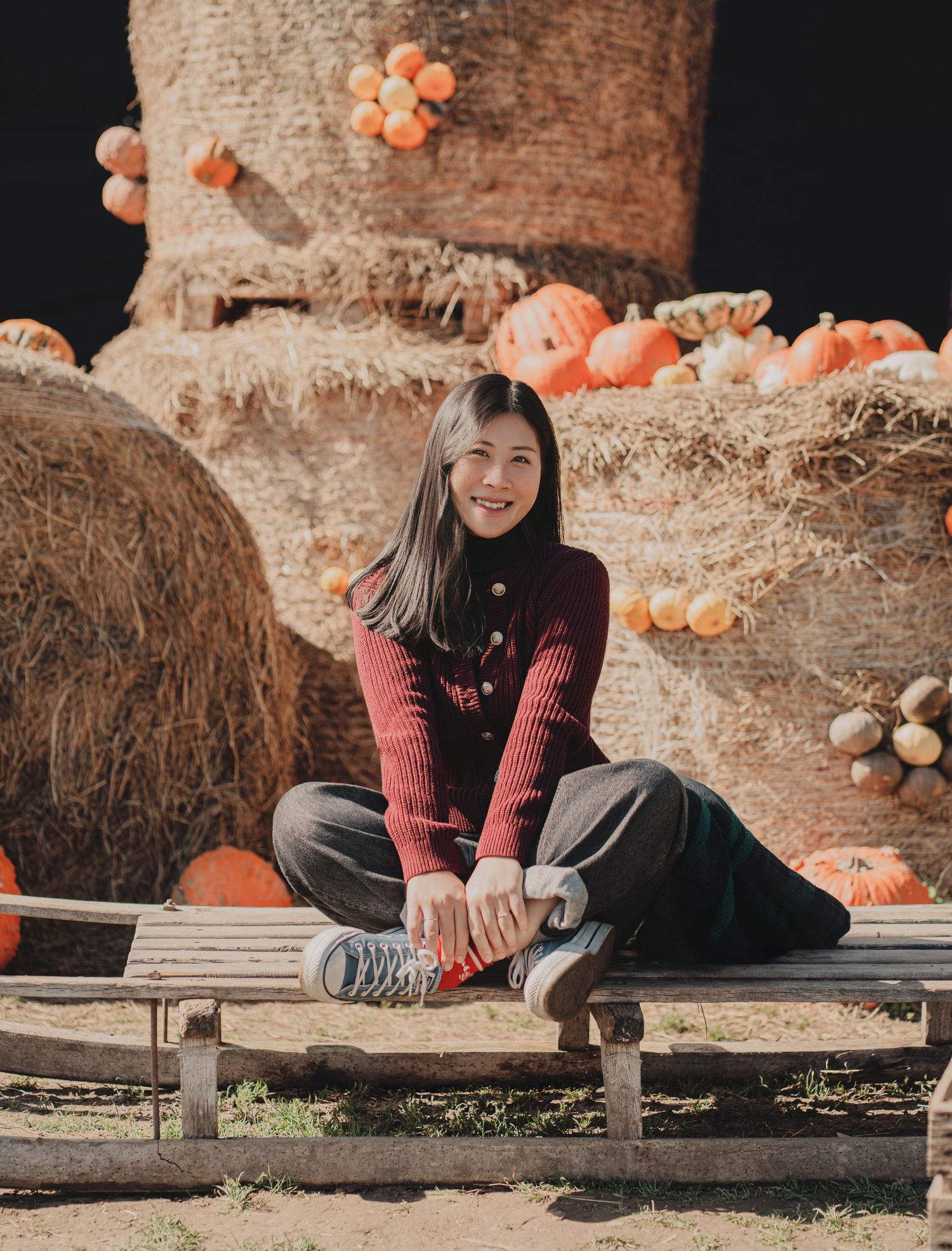 Young woman in dark red knit sweater smiles while sitting on wooden bench at sunny fall pumpkin patch.