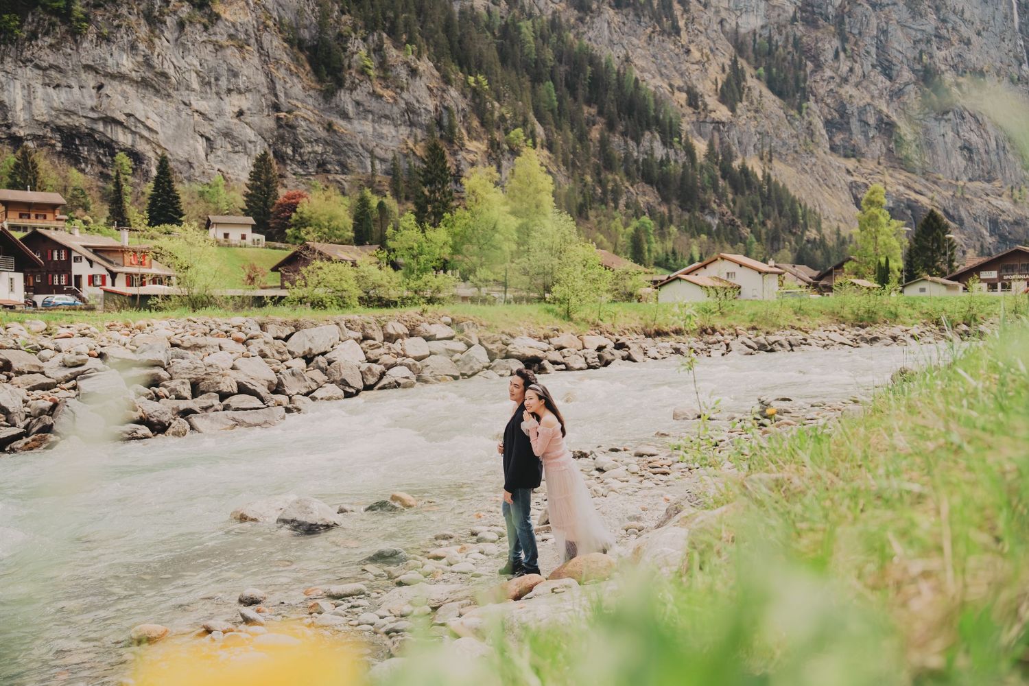 Couple sharing a tender embrace on rocky riverbank with Swiss alpine village and mountains behind.