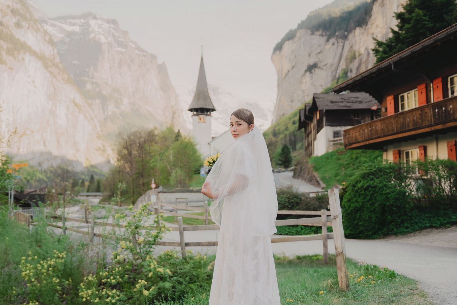Bride in white lace gown with veil holds yellow flowers near a Swiss alpine church.
