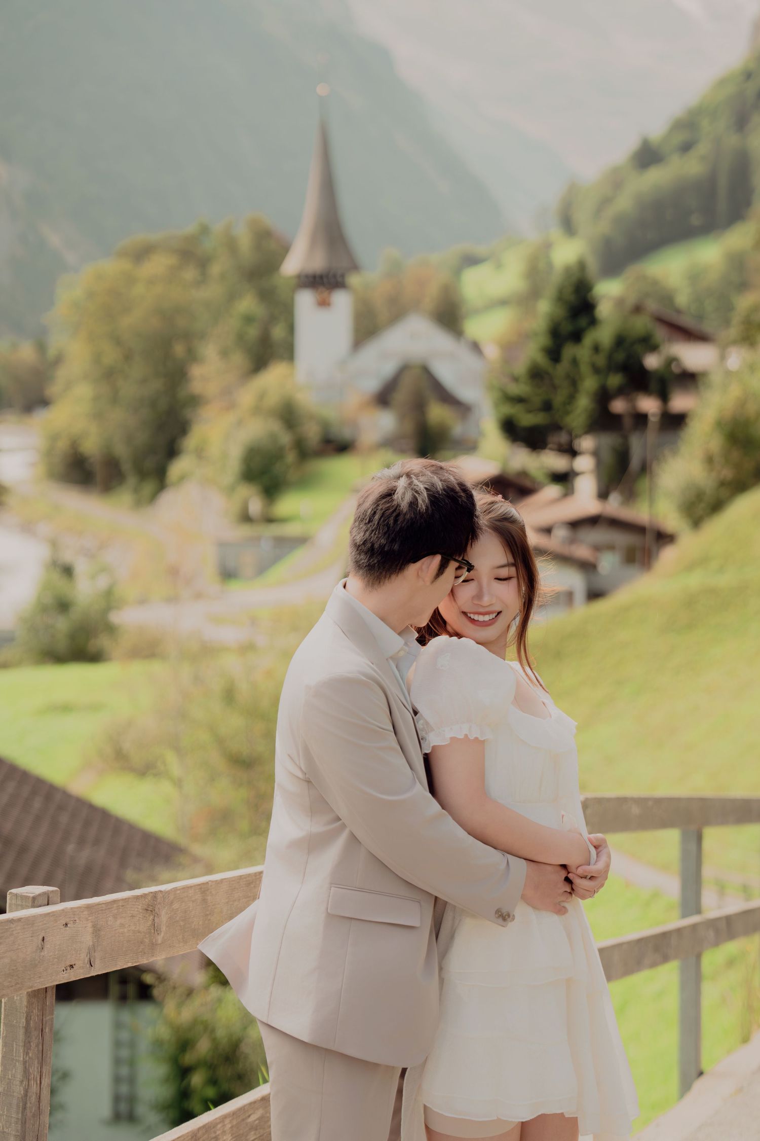 Couple embracing tenderly on wooden bridge with Swiss church steeple and alpine village in background.