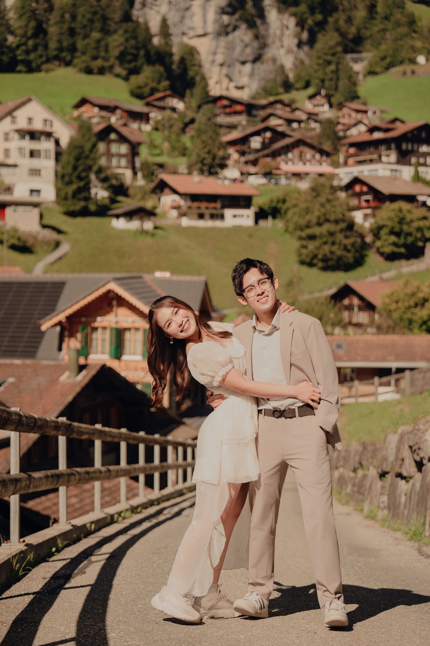 Couple posing and smiling on a road with traditional Swiss chalets on a hillside.