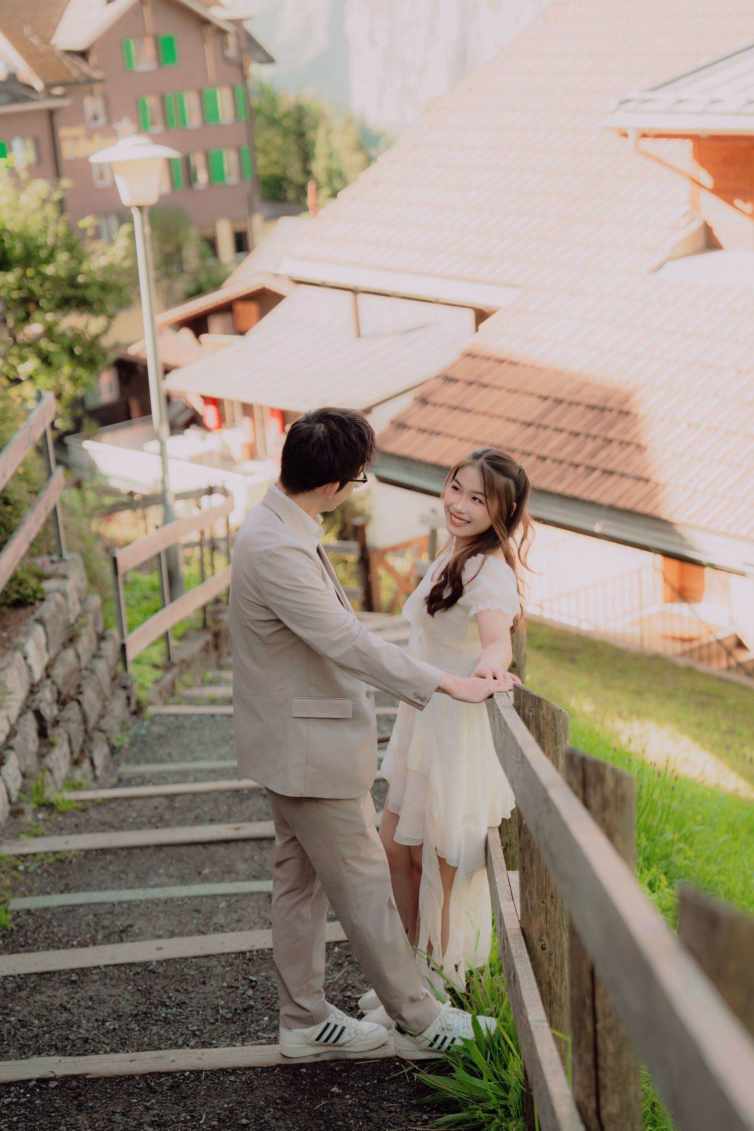 Couple holding hands on scenic Swiss village staircase, woman in white dress smiling, alpine buildings in background.