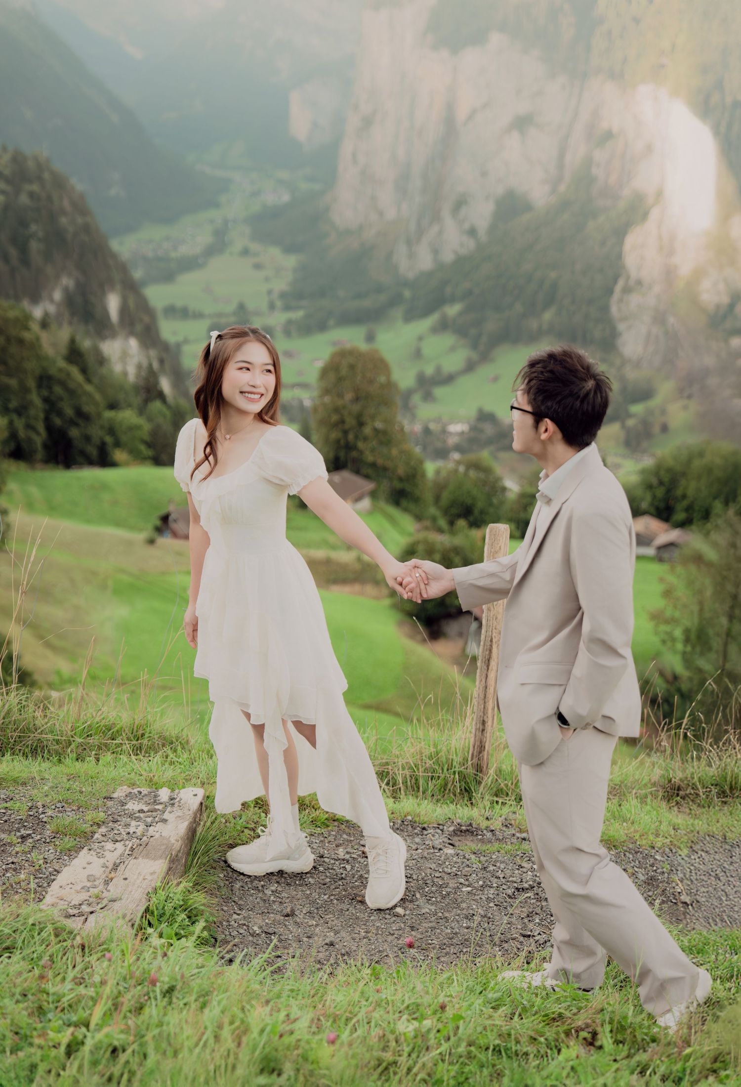 Smiling bride in white dress holding hands with groom in beige suit on a scenic Swiss mountain hillside.
