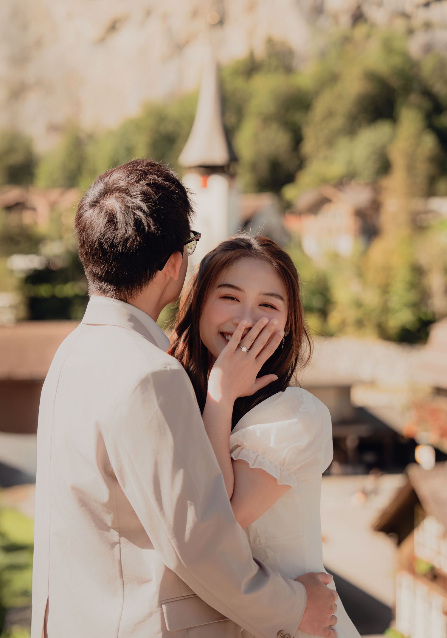 Bride laughing joyfully with hand over mouth embraced by groom near Swiss village church.