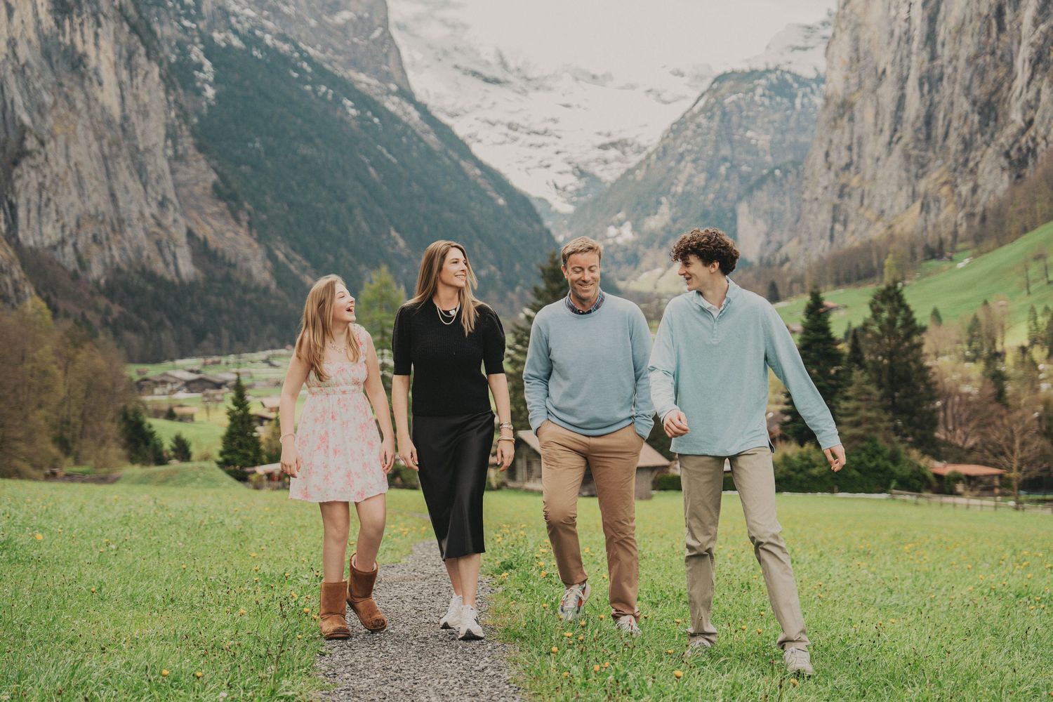Happy family of four walking hand-in-hand on green meadow path in Swiss Alps valley.