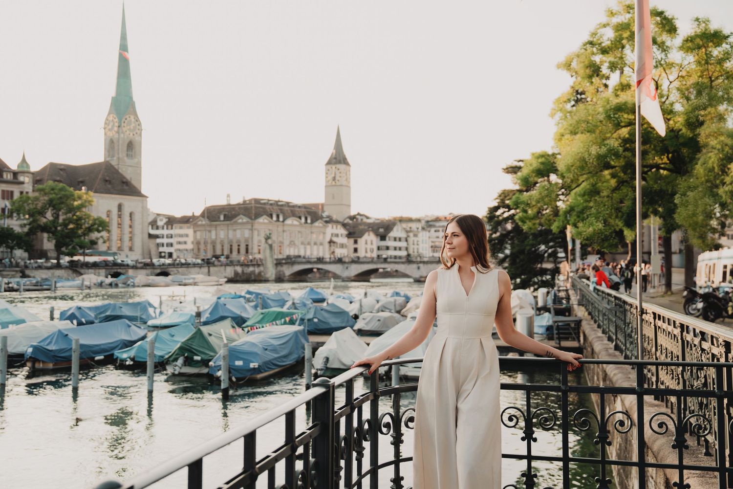 Woman in white dress poses on a bridge along Zurich's Limmat River with historic church spires and boats in background.