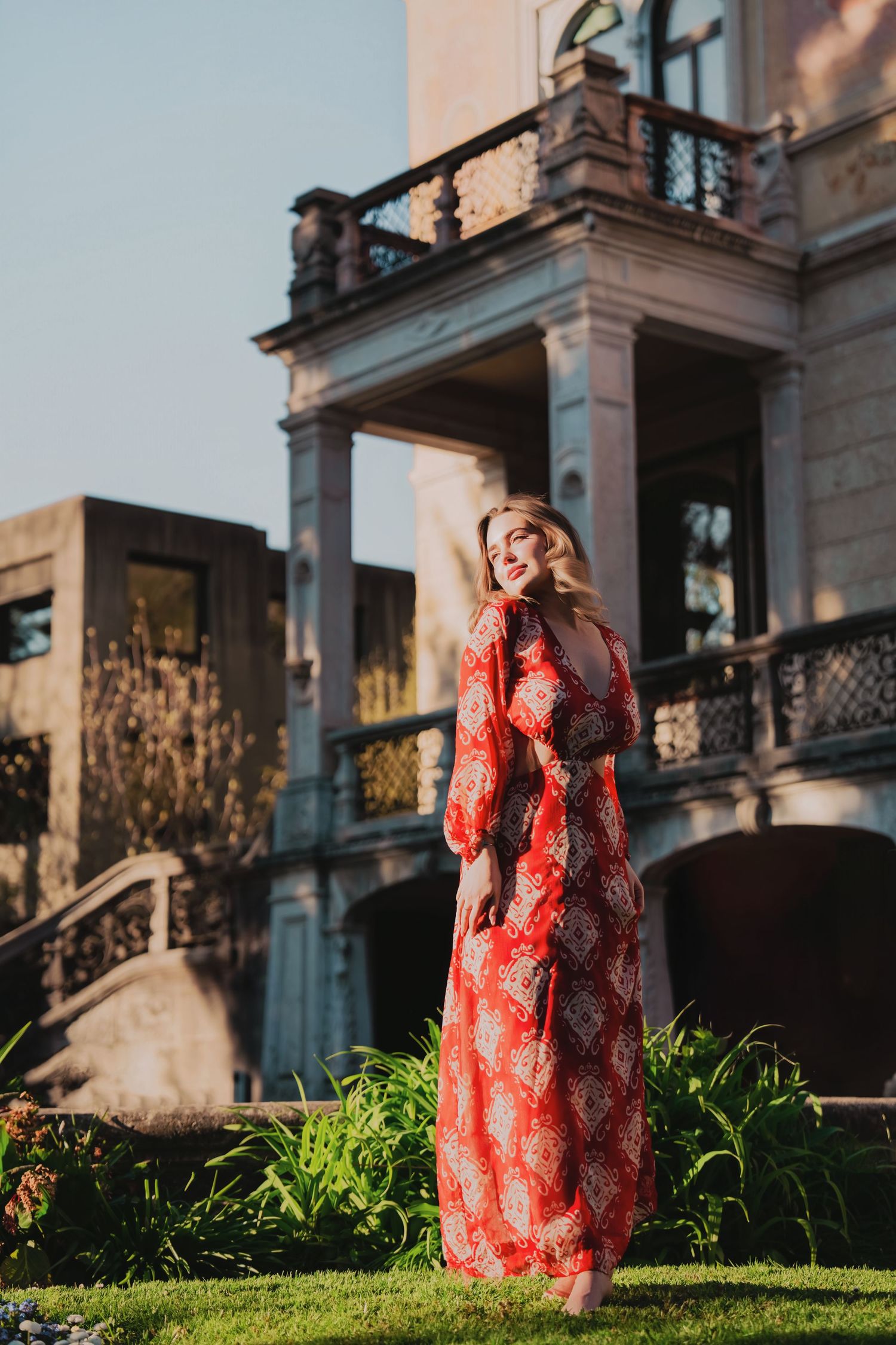 Woman in red floral maxi dress poses smiling in front of a grand historic building with balconies.