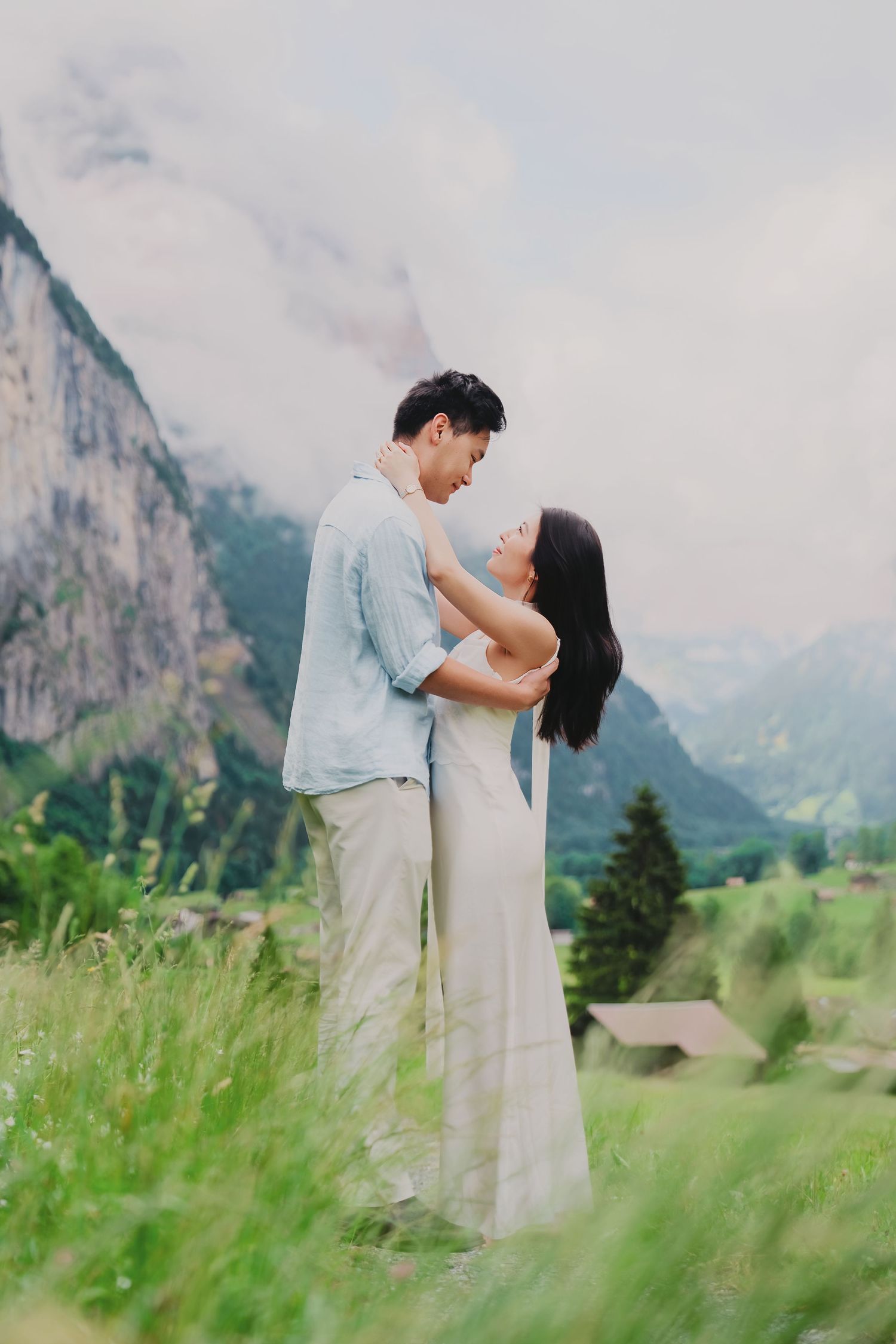Couple embracing in a lush green alpine meadow with dramatic misty mountains in the background.