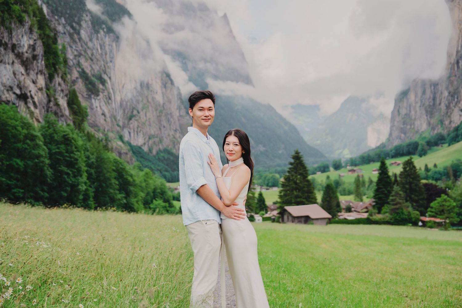 Couple poses together in green Swiss alpine meadow with dramatic misty mountain cliffs and valley views.