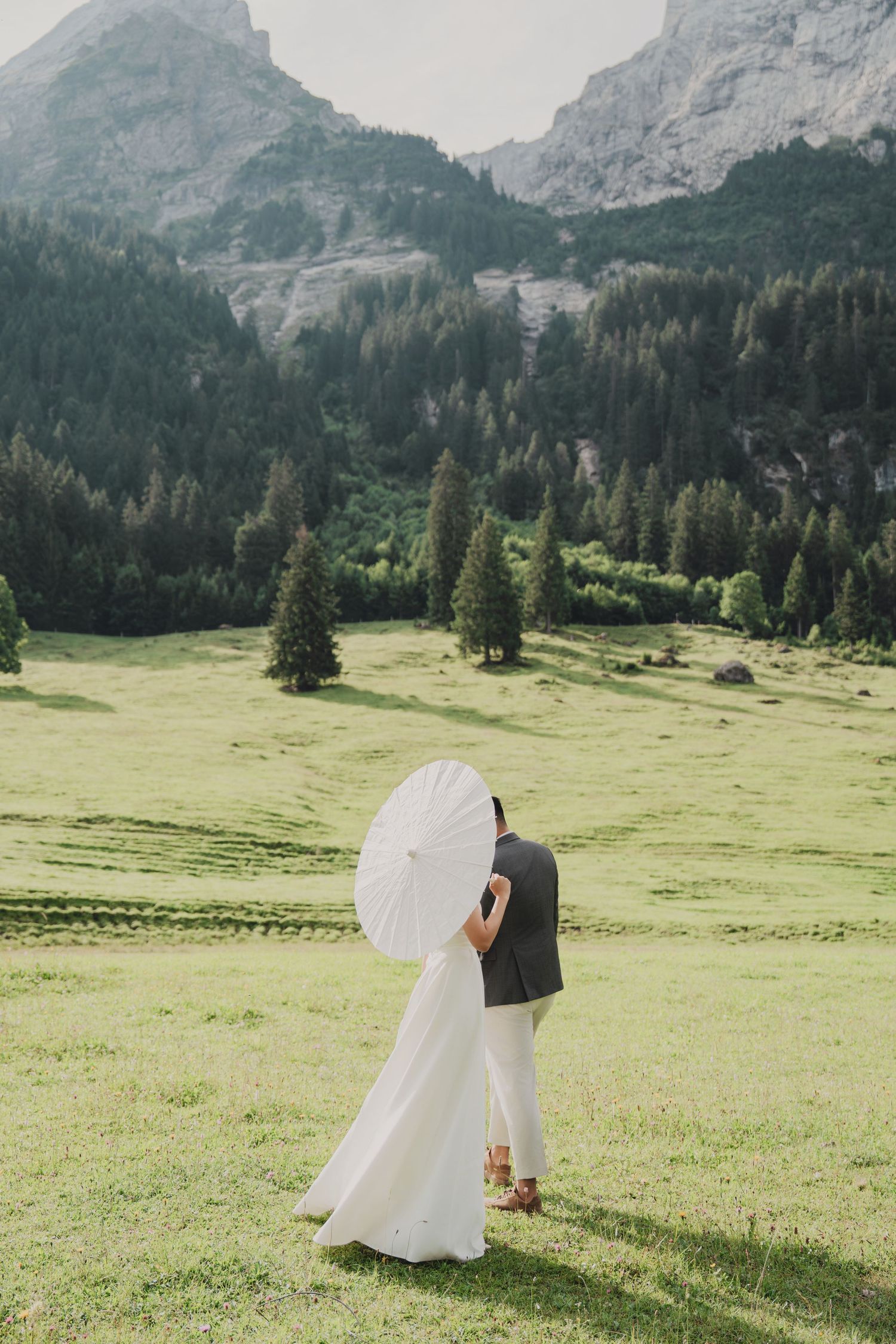Wedding couple embraces behind white parasol in green alpine meadow with dramatic rocky mountain peaks in background.