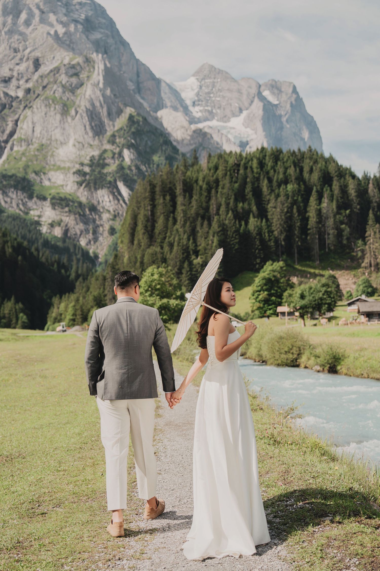 Bride with white parasol and groom walking along river path with alpine mountains in background.