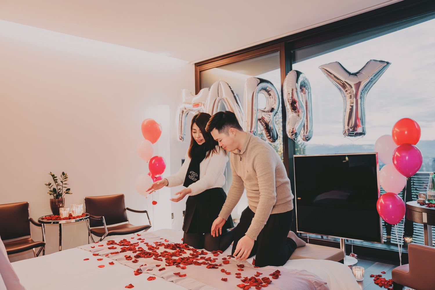 Couple arranging rose petals on bed in romantic hotel room decorated with balloons and flowers.