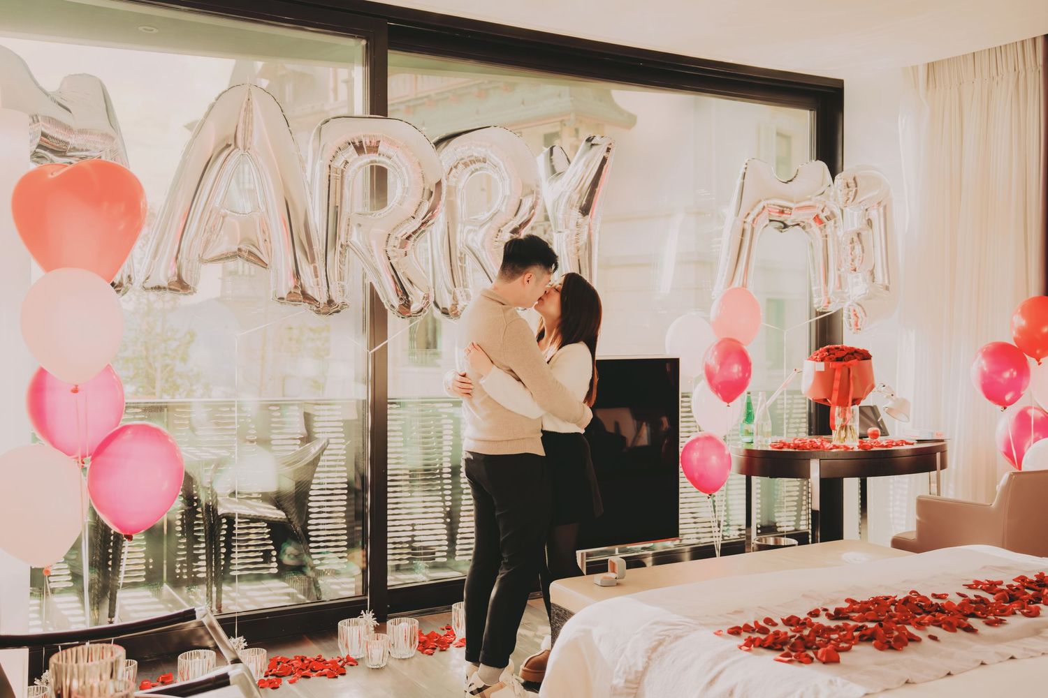 Joyful couple embracing after a successful marriage proposal surrounded by MARRY balloons and colorful heart balloons.