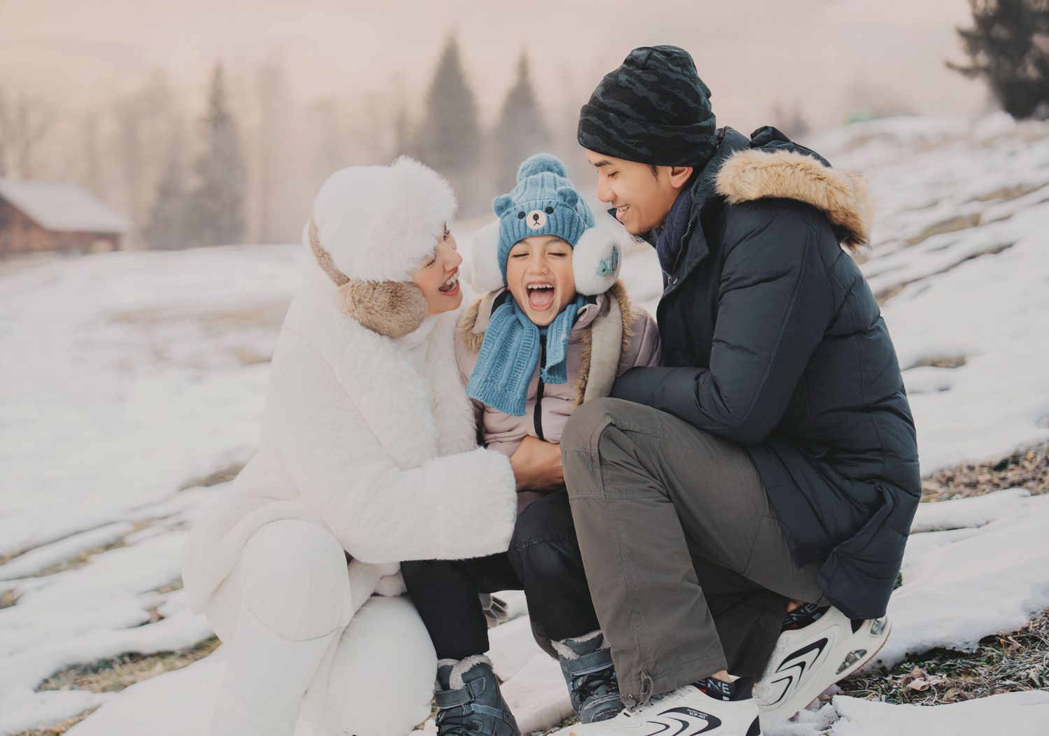 Family of three laughing together in the snow, child in blue hat between parents in winter clothing.