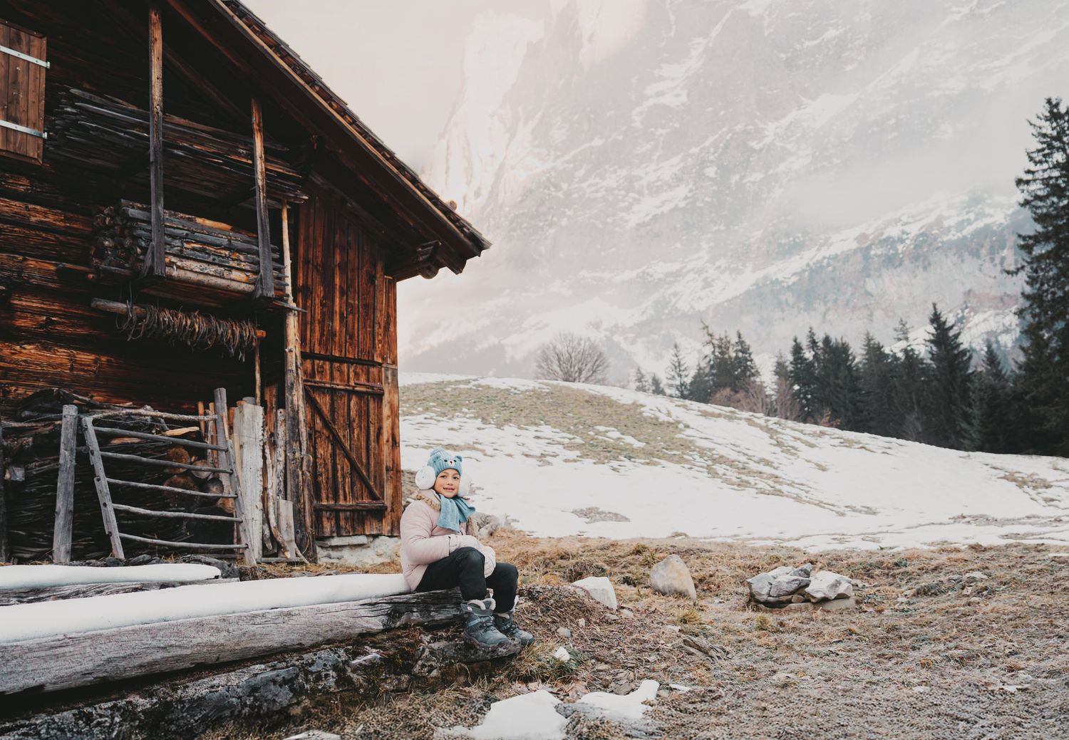 Child in blue hat sitting alone beside rustic wooden alpine cabin with snowy mountain backdrop.