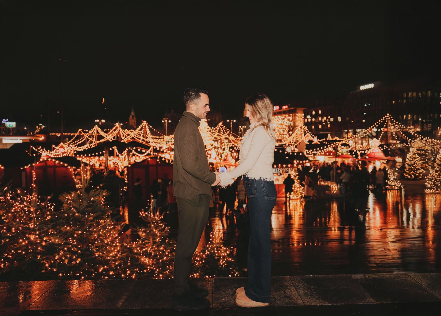 Couple facing each other at illuminated Christmas market with warm festive lights.