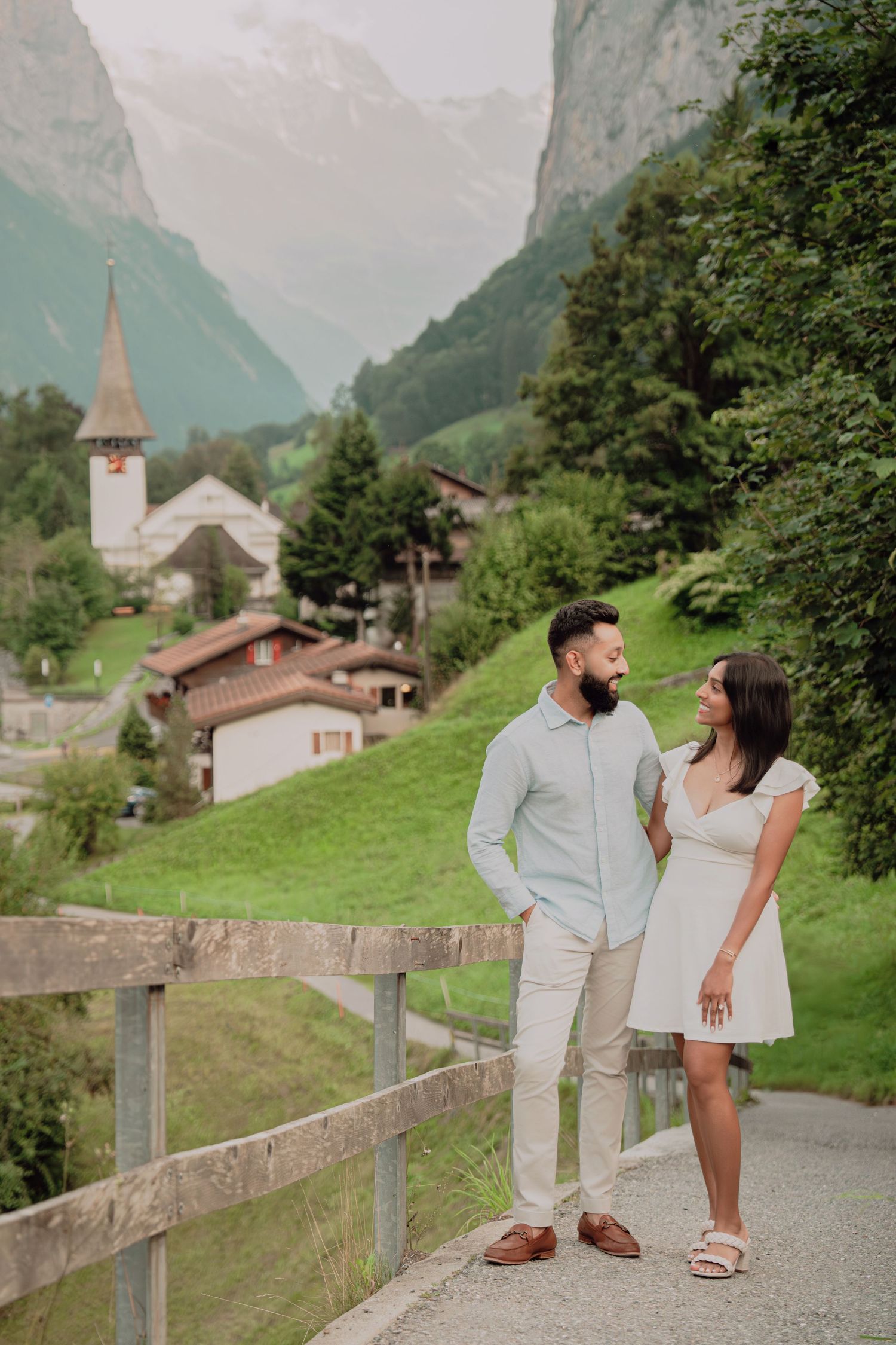 Couple standing together on wooden bridge path with scenic Swiss village church and towering mountain backdrop behind them.