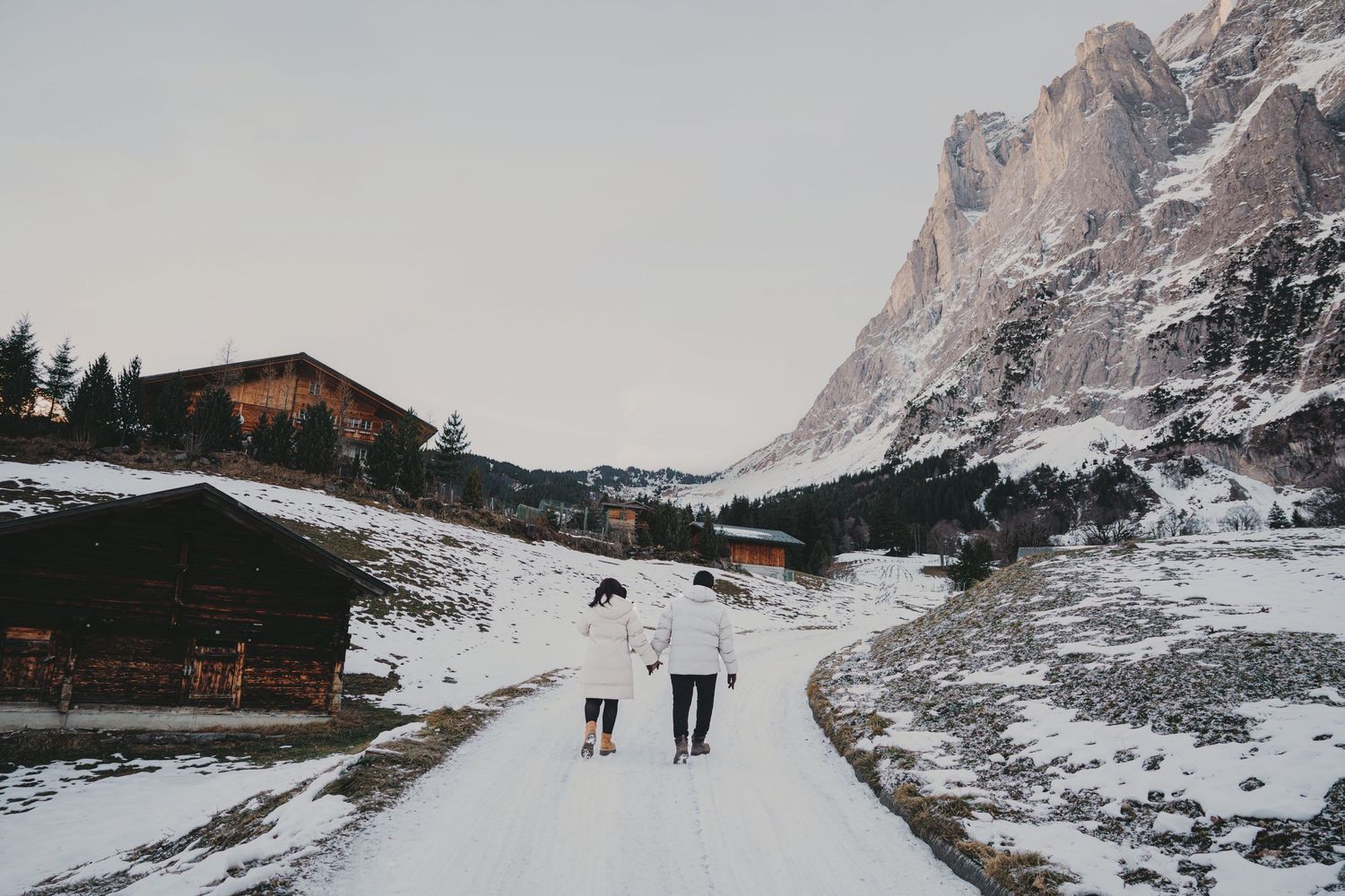 Couple walking hand in hand down snowy path between wooden chalets toward mountain scenery.