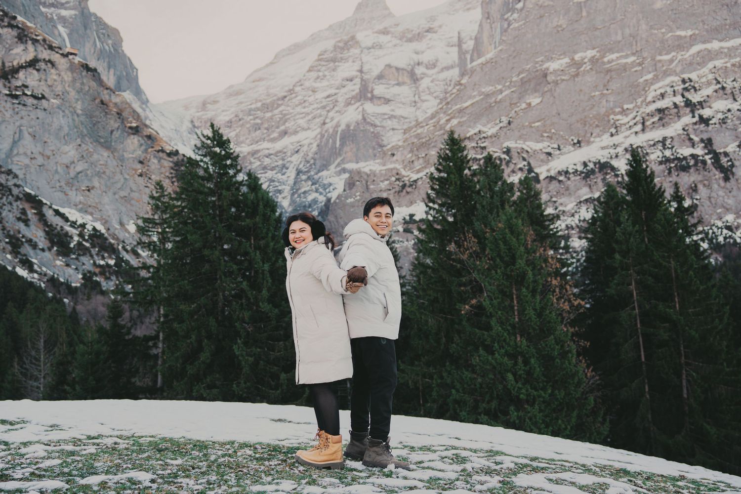 Couple standing back-to-back holding warm drinks with dramatic snowy mountain backdrop.