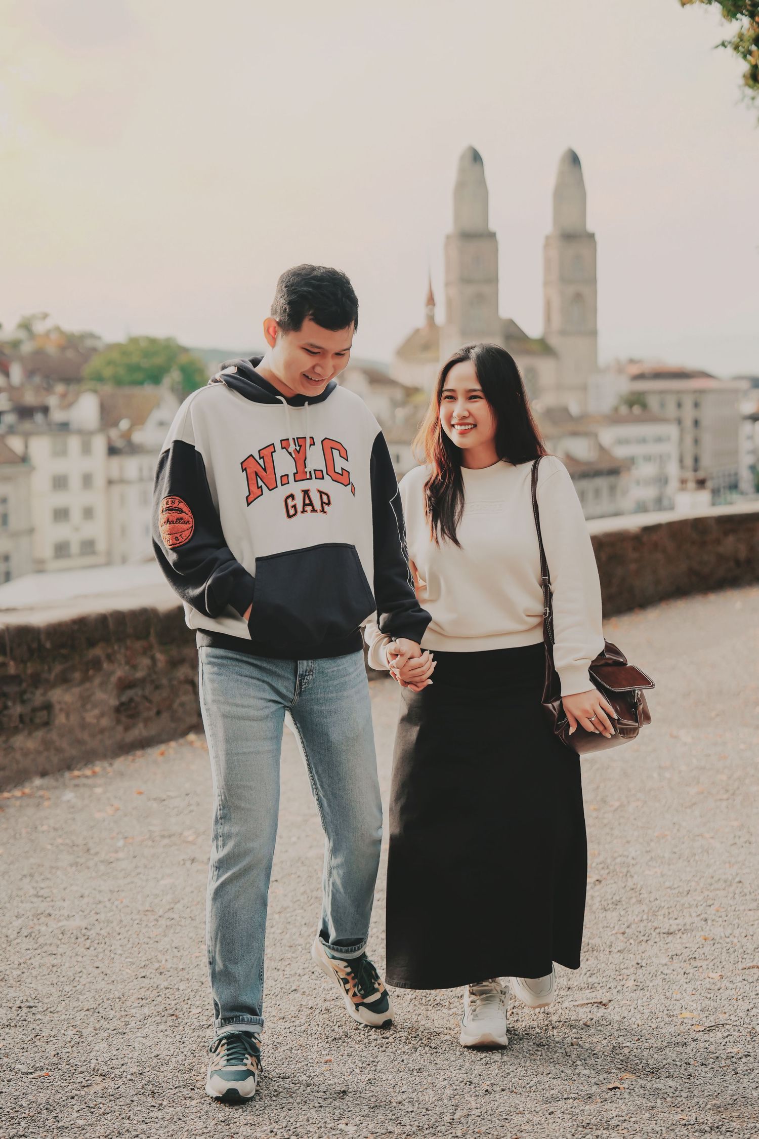 A laughing couple walks hand-in-hand on a terrace with Zurich's Grossmünster cathedral towers in the background.