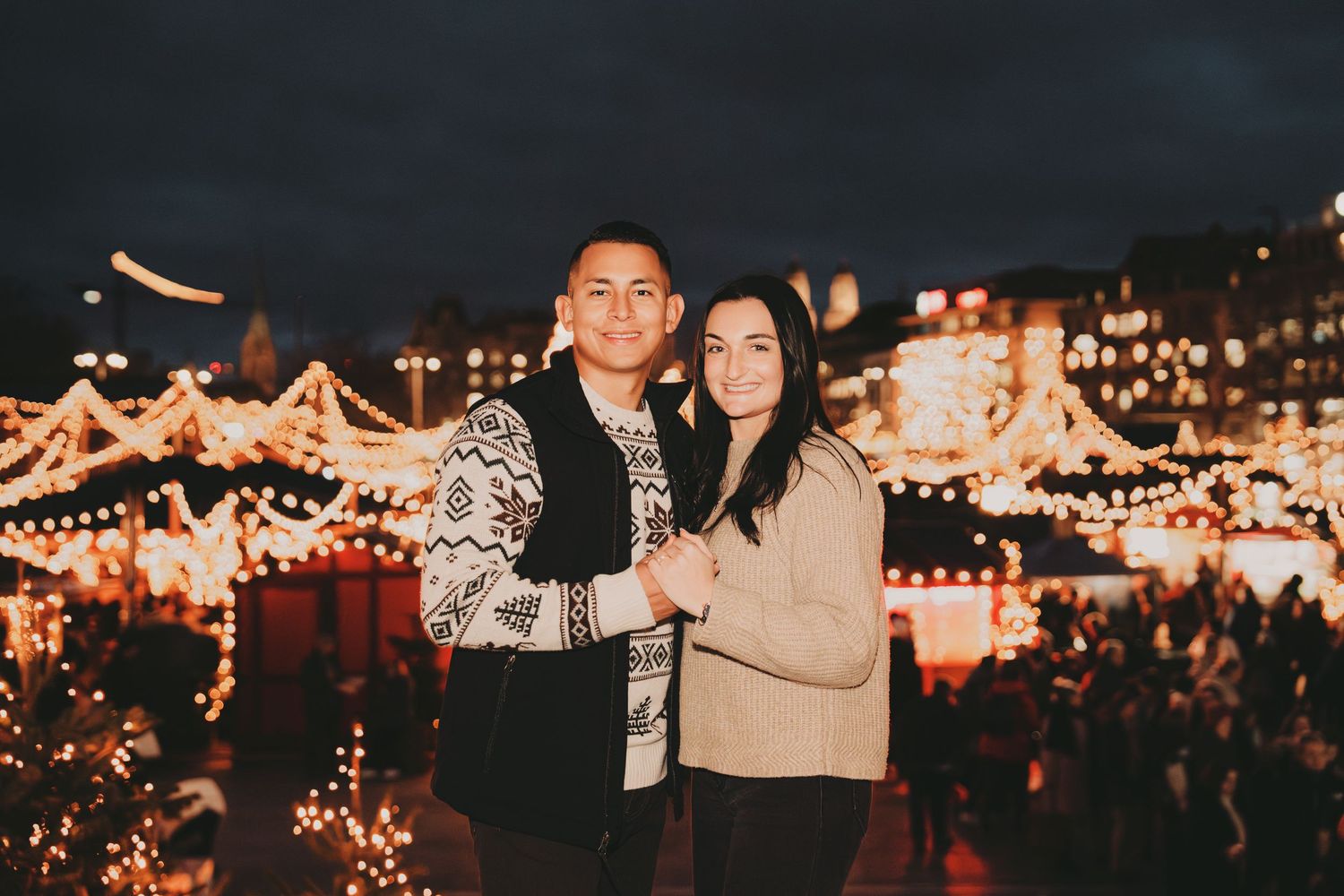 Smiling couple posing together at a beautifully lit Christmas market with golden holiday decorations.