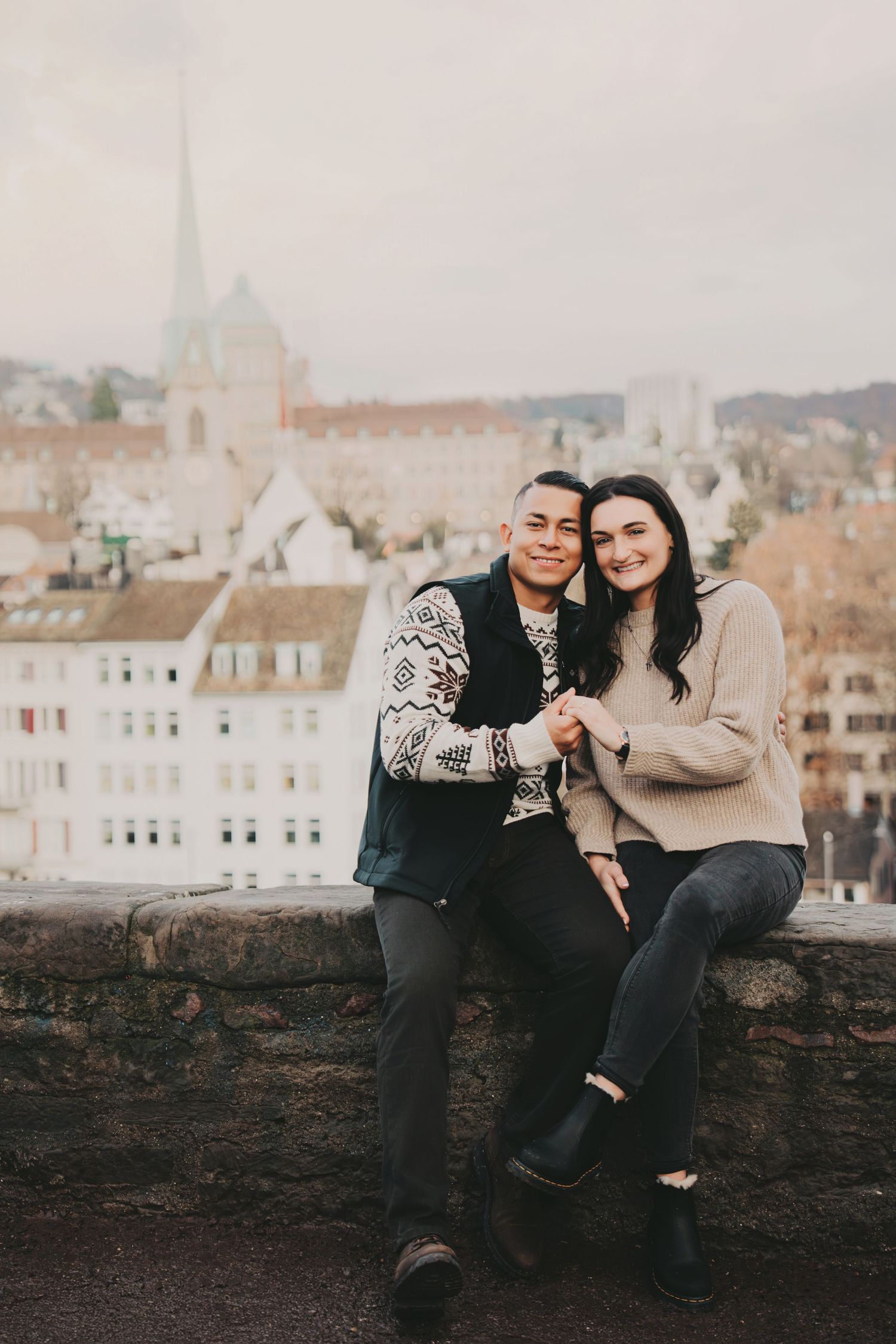 Happy couple sitting together on a stone wall with a scenic European city backdrop.