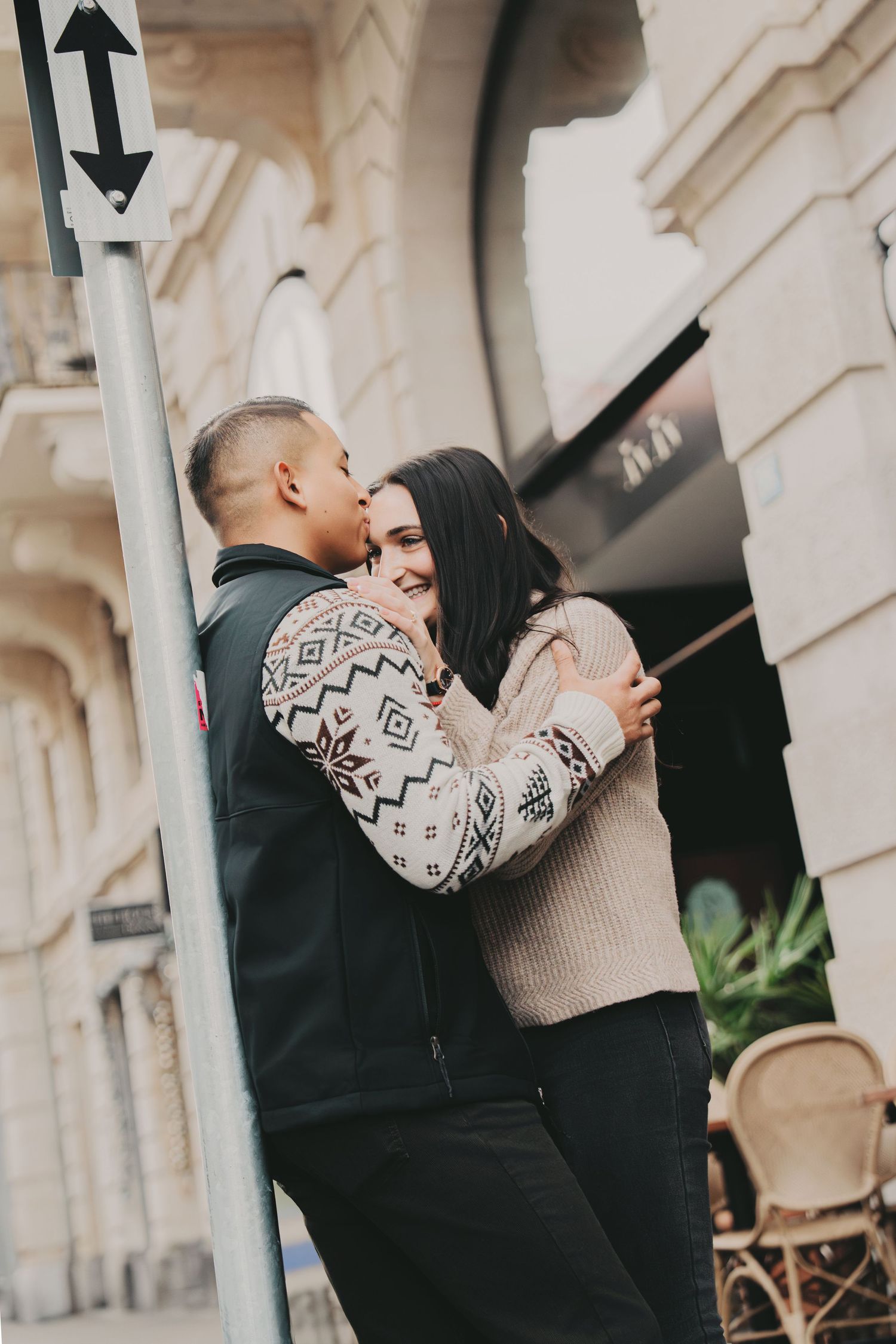 Man kissing womans cheek while embracing outside a classic European building storefront.
