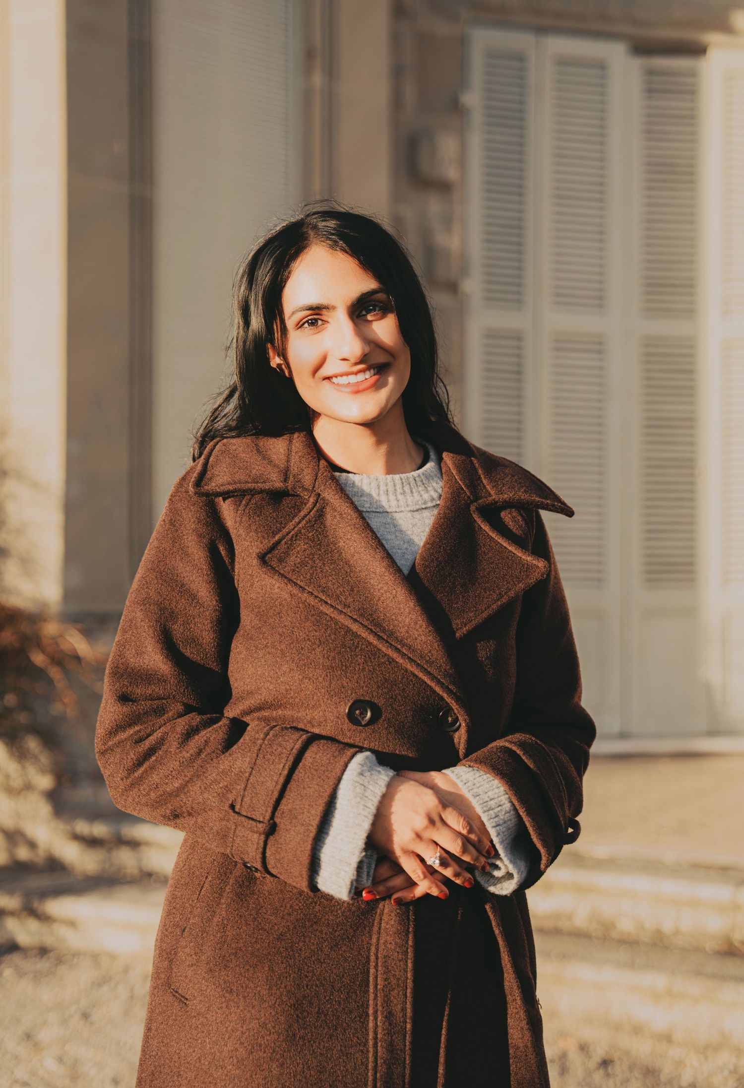 Smiling South Asian woman in brown wool coat standing in warm sunlight outside a building.