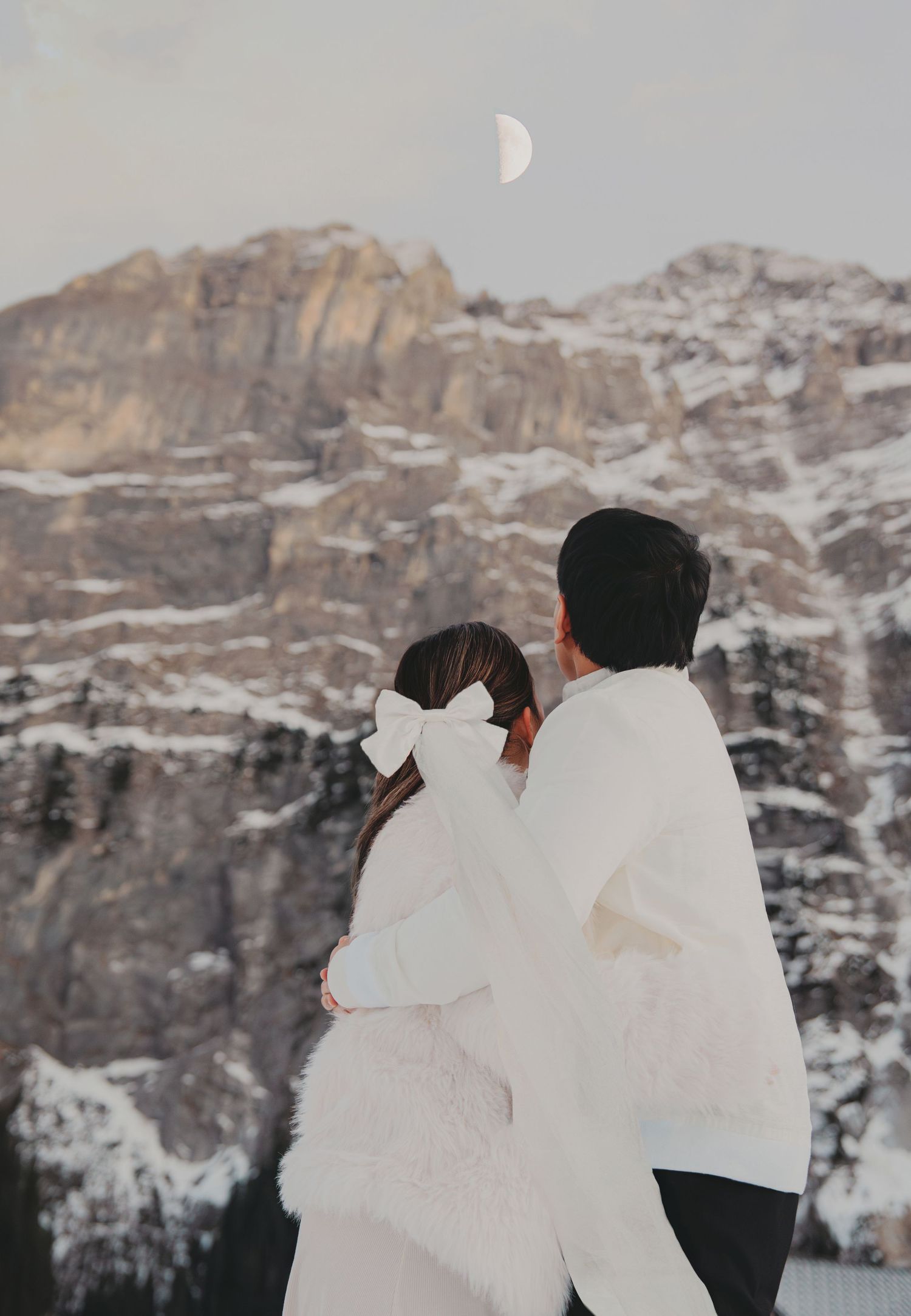 Couple embracing from behind in white winter outfits gazing at snowy mountain with crescent moon.