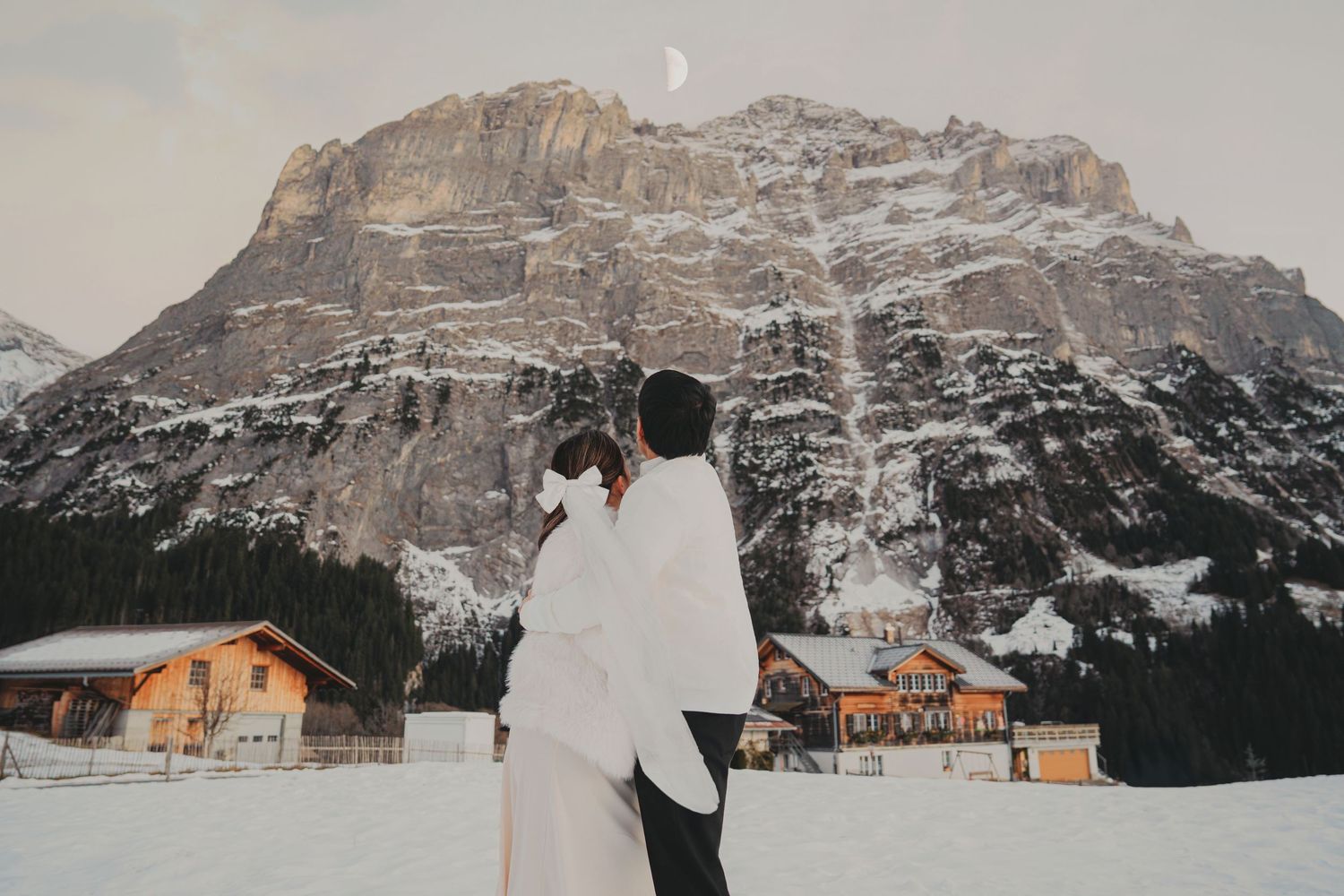 Romantic couple embracing in snow with crescent moon over Alpine mountain village.