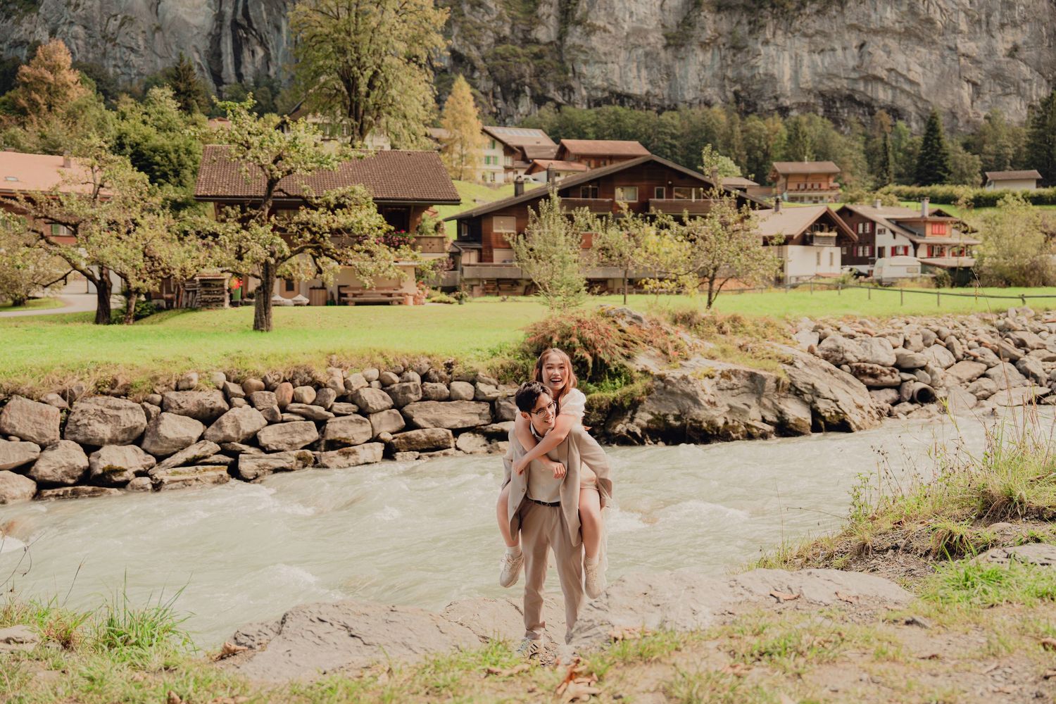 Man giving woman a piggyback ride beside a rushing river with Swiss alpine scenery in Lauterbrunnen.