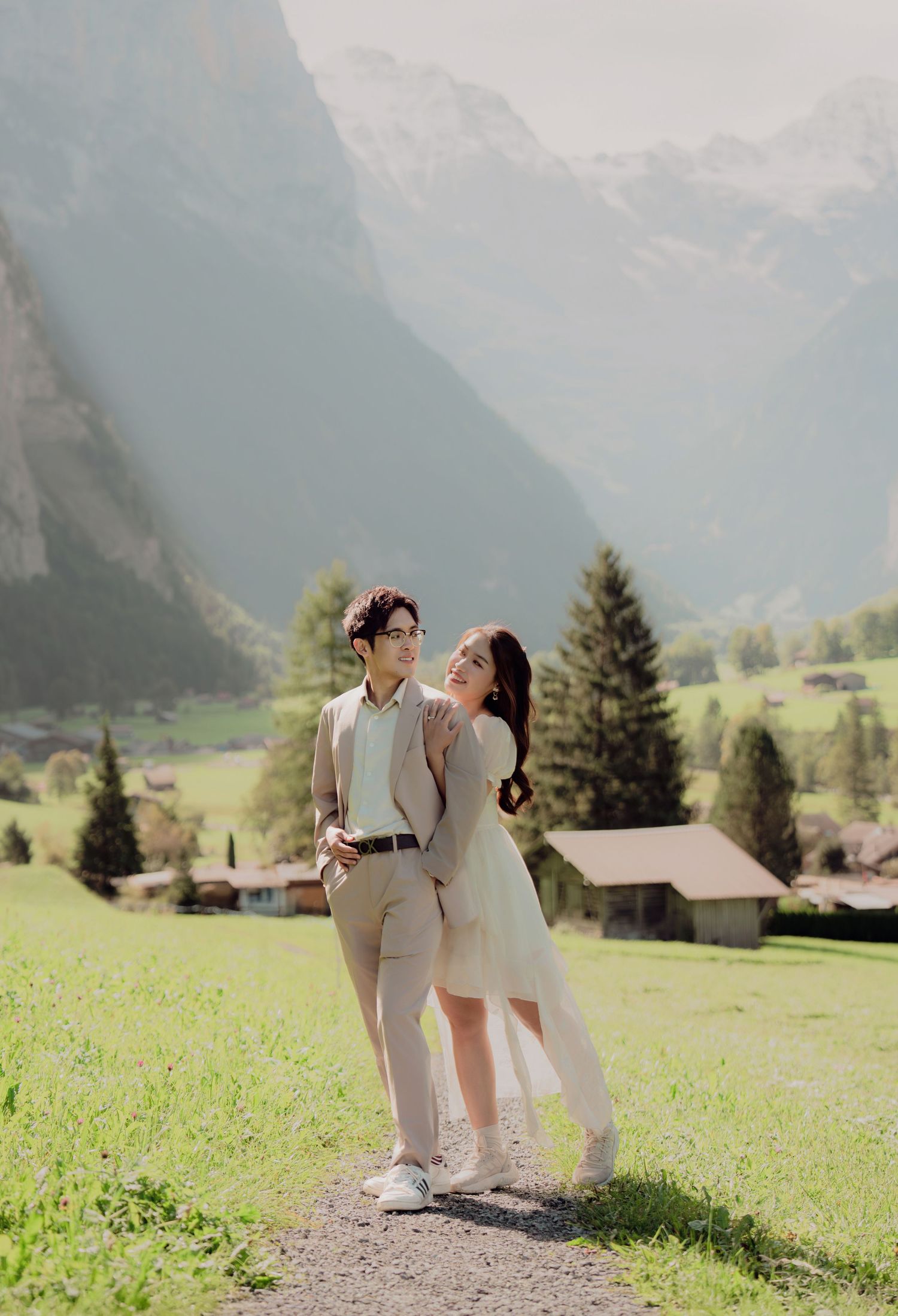 Couple in coordinating beige wedding outfits embracing on a sunlit path with Swiss Alpine scenery behind them.