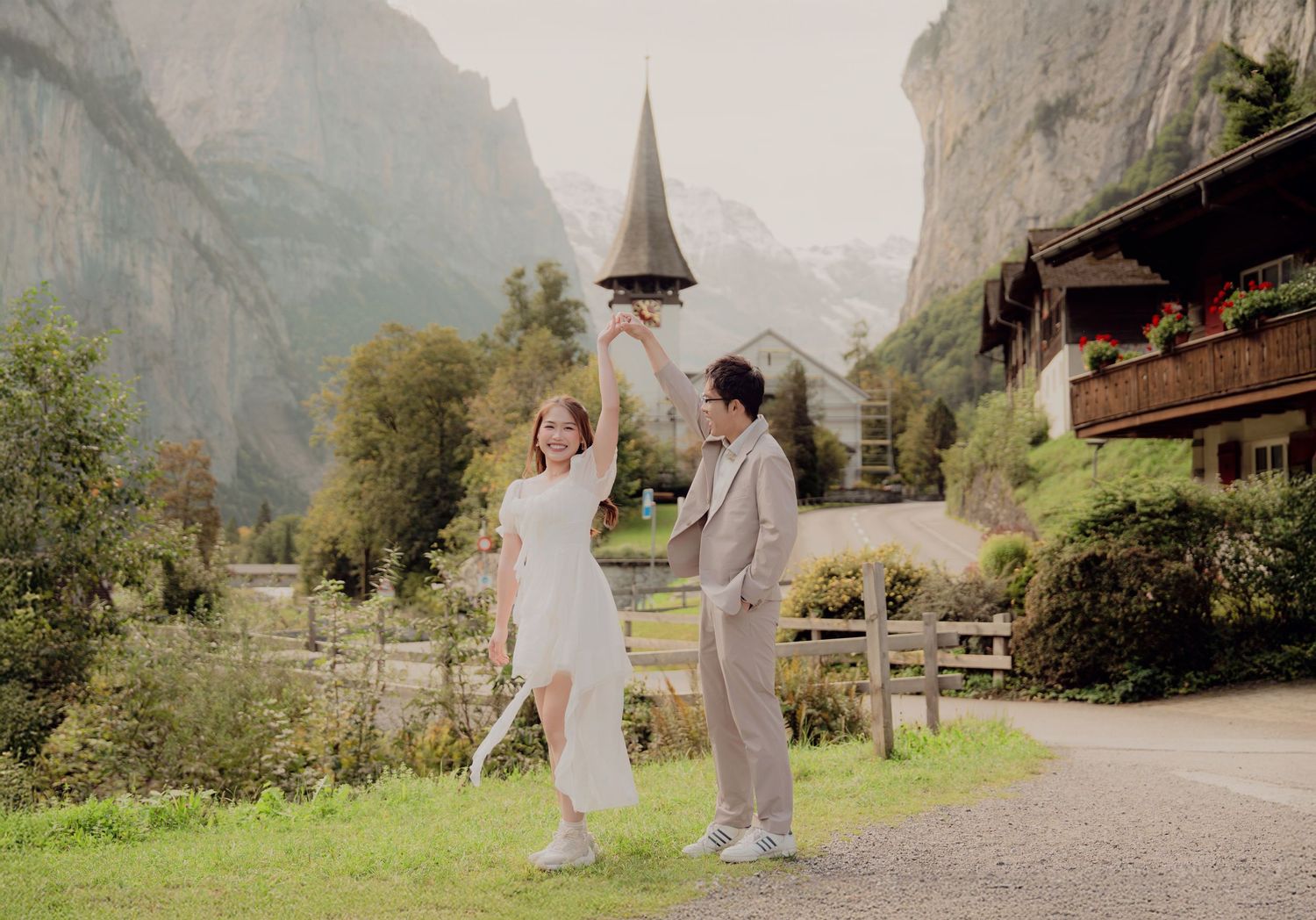 Couple dancing playfully on a path with a Swiss church steeple and mountains behind.