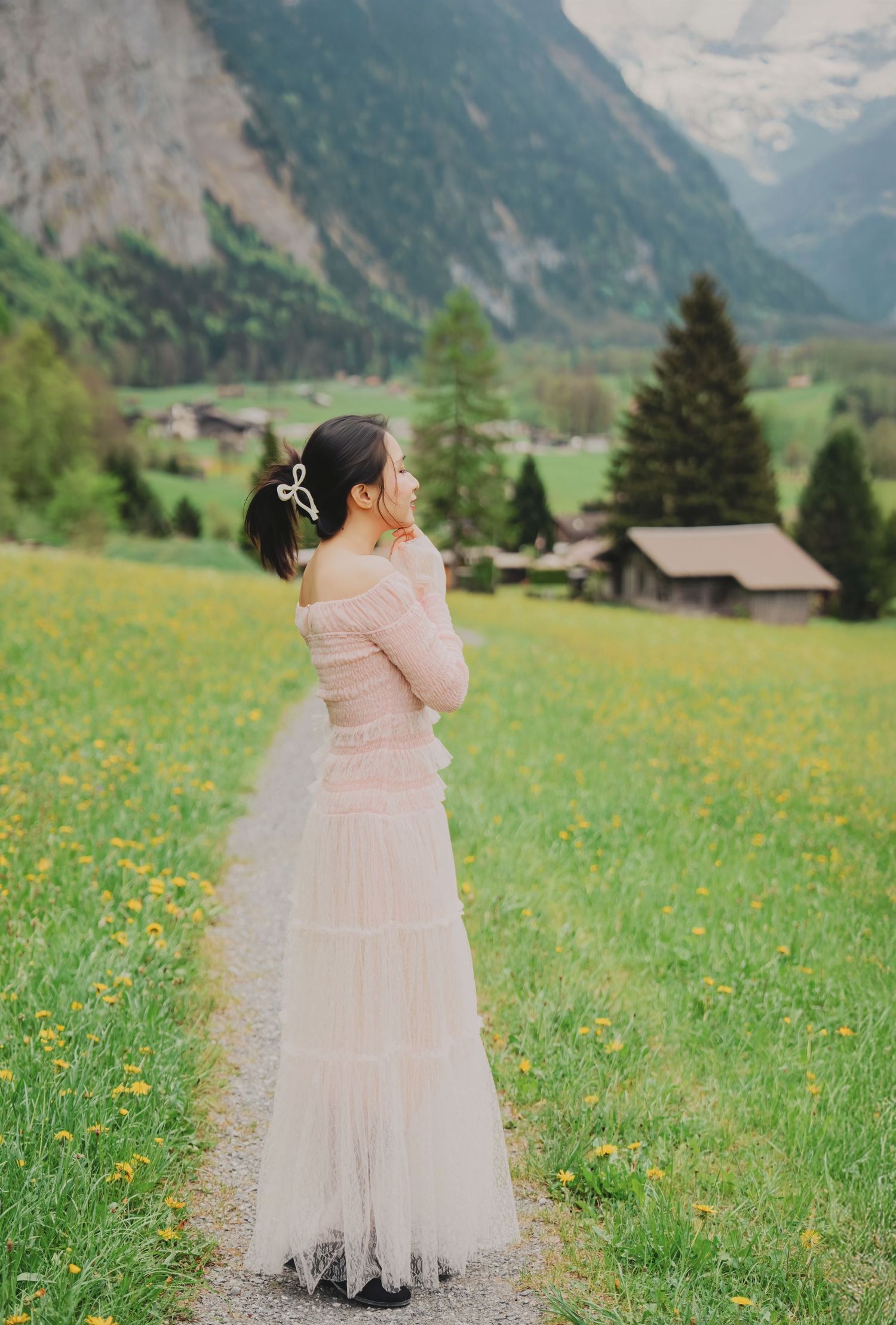 Woman in off-shoulder pink dress standing in Swiss alpine meadow with yellow wildflowers and mountains.