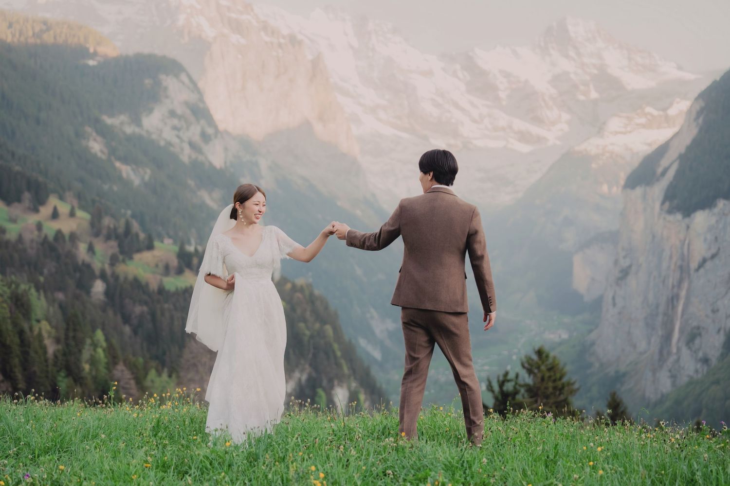 Bride and groom walking hand in hand across a lush green alpine meadow with dramatic Swiss mountain scenery.