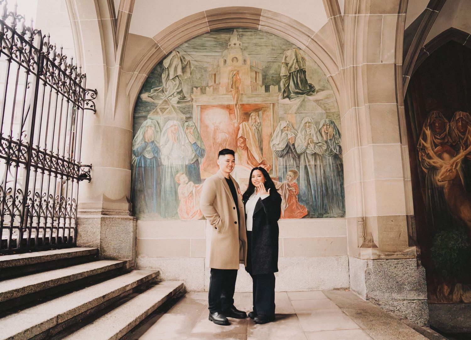 Engaged couple standing together on cathedral steps in front of a large religious fresco.