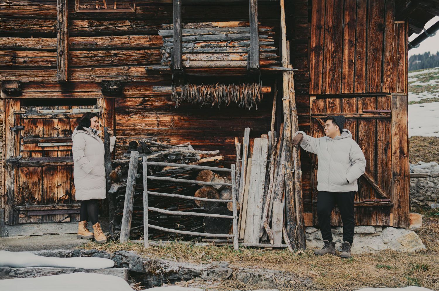 Couple in white winter coats exploring rustic alpine wooden barn with dried herbs hanging outside.