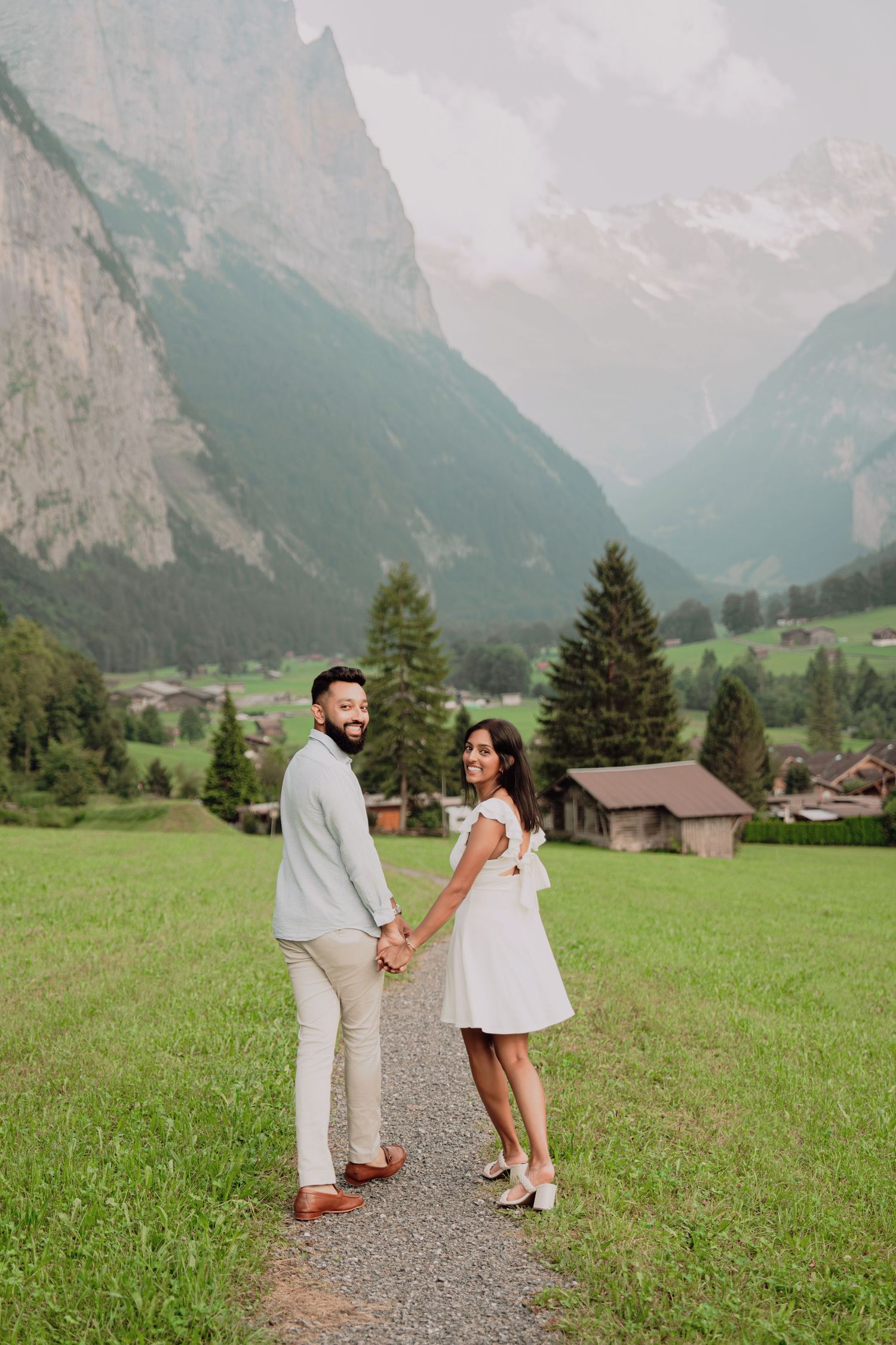Couple holding hands on a path, looking back and smiling, with a stunning Swiss Alpine valley backdrop.