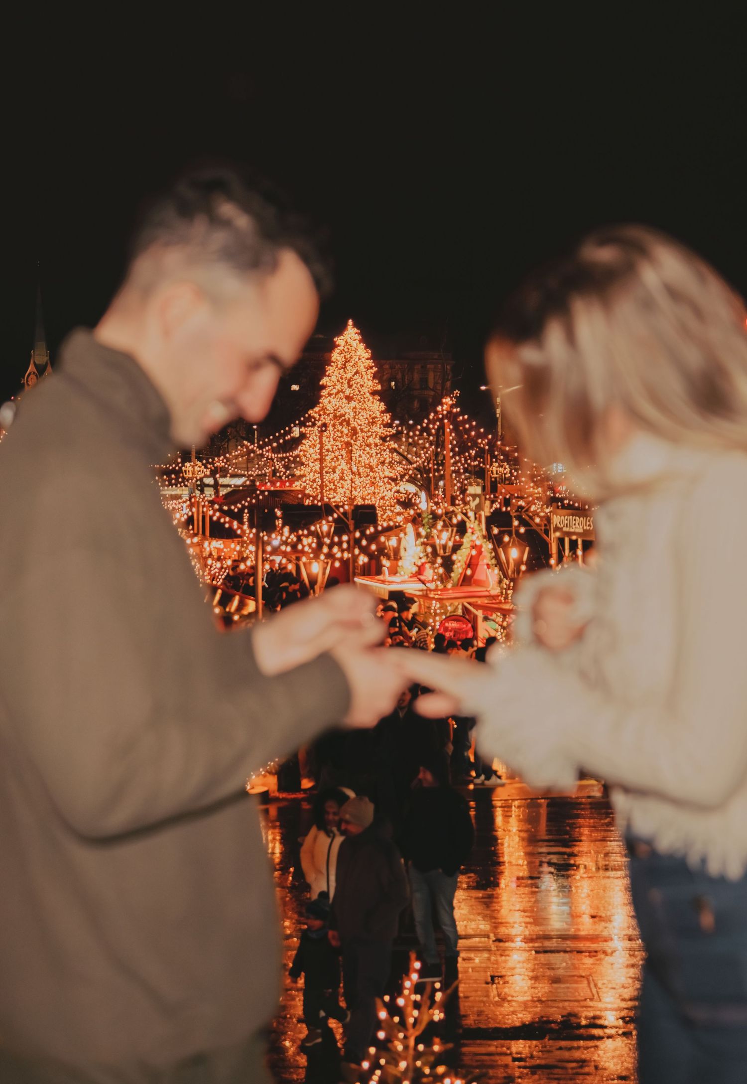 Couple holding hands at a Christmas market with a glowing decorated tree and festive lights behind them.