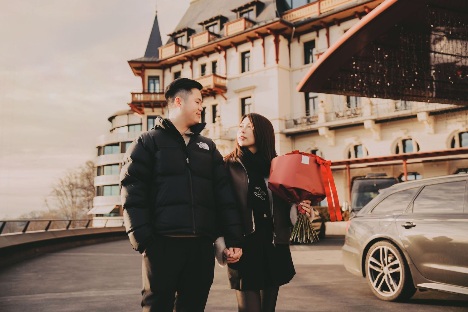 Asian couple holding hands and smiling at each other outside a grand hotel with a red gift bag.