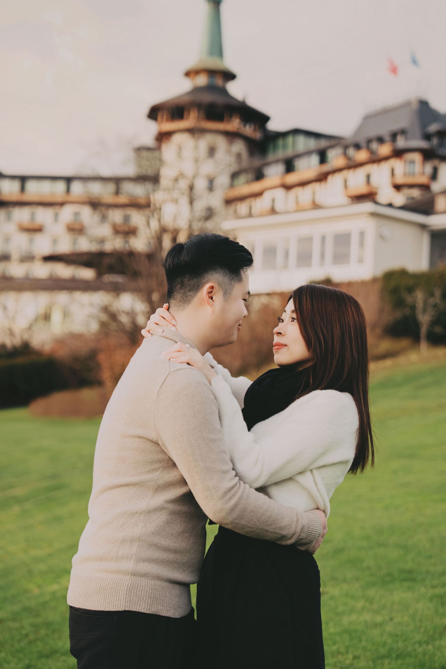 Young Asian couple embracing romantically on green lawn outside castle-style hotel.