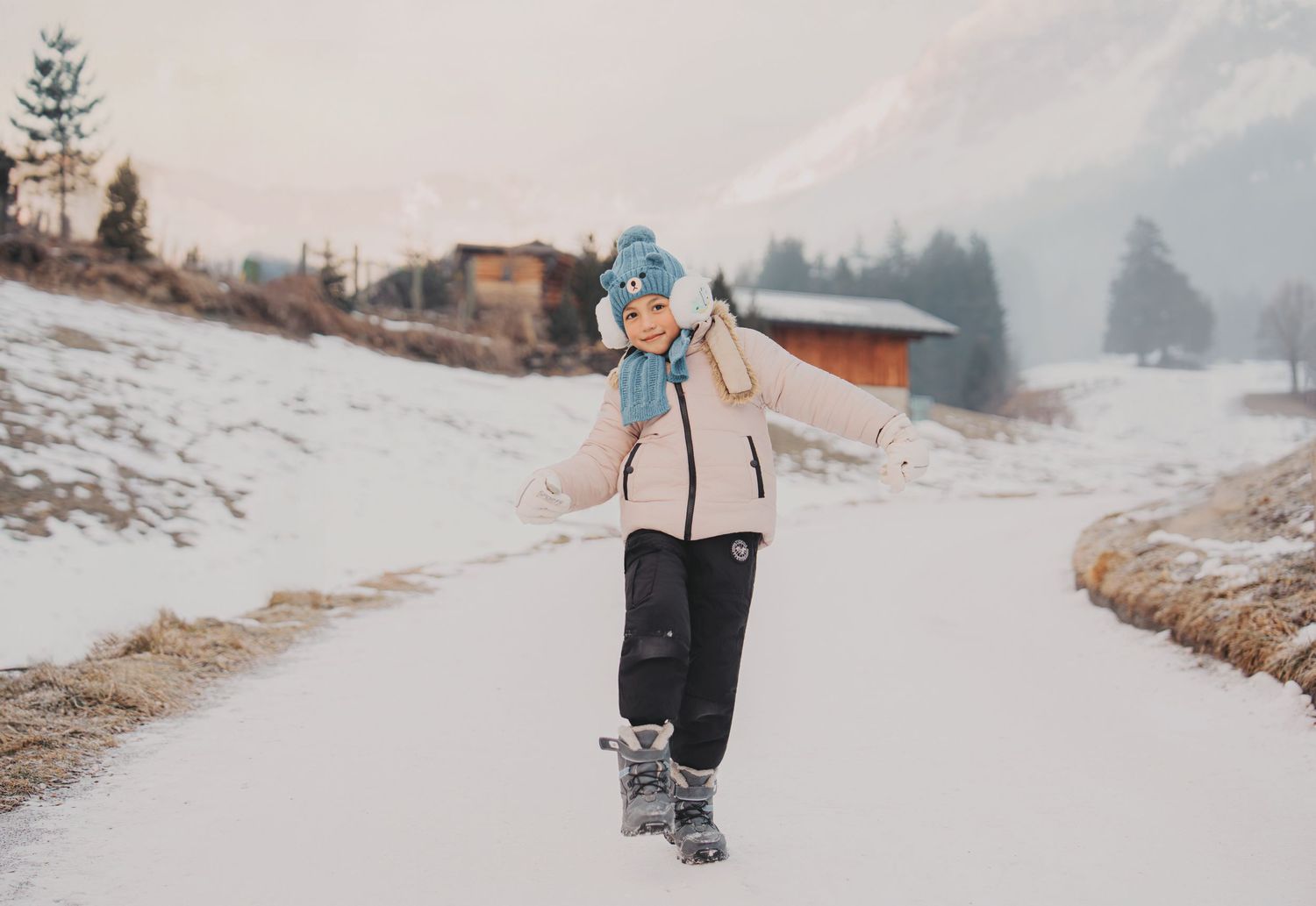 Young child in blue beanie and scarf walking on snowy alpine road with wooden chalets and pine trees.