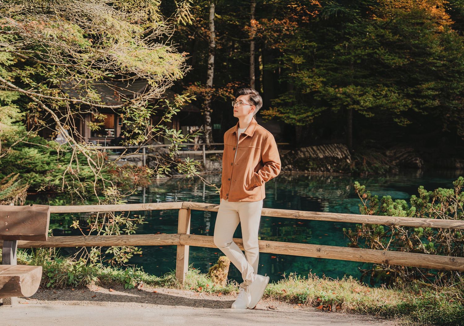 Young man in brown jacket and white pants stands by wooden fence near a tranquil blue lake in autumn forest.