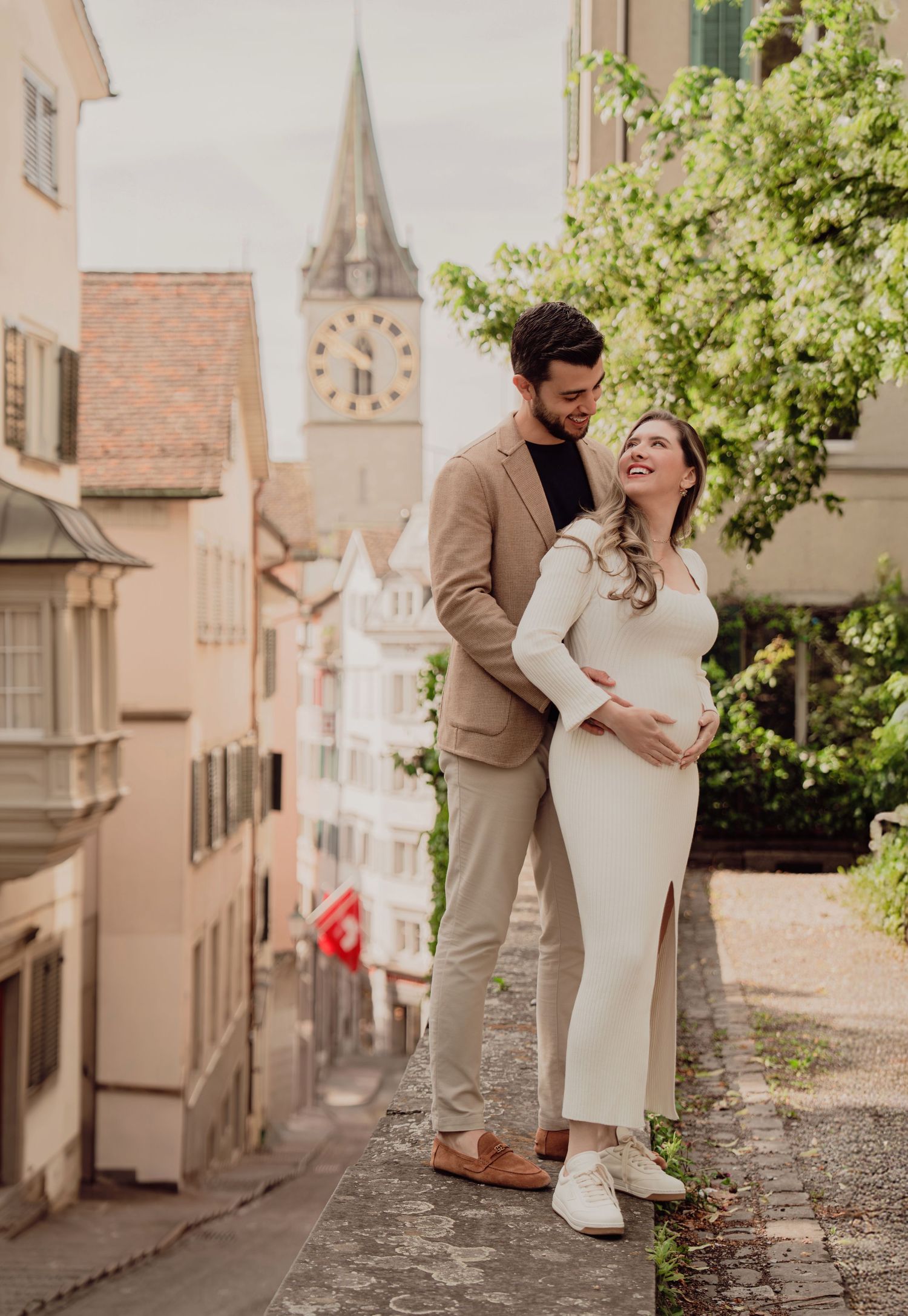 Happy expecting couple laughs together during maternity shoot with St. Peter church tower in Zurich background.