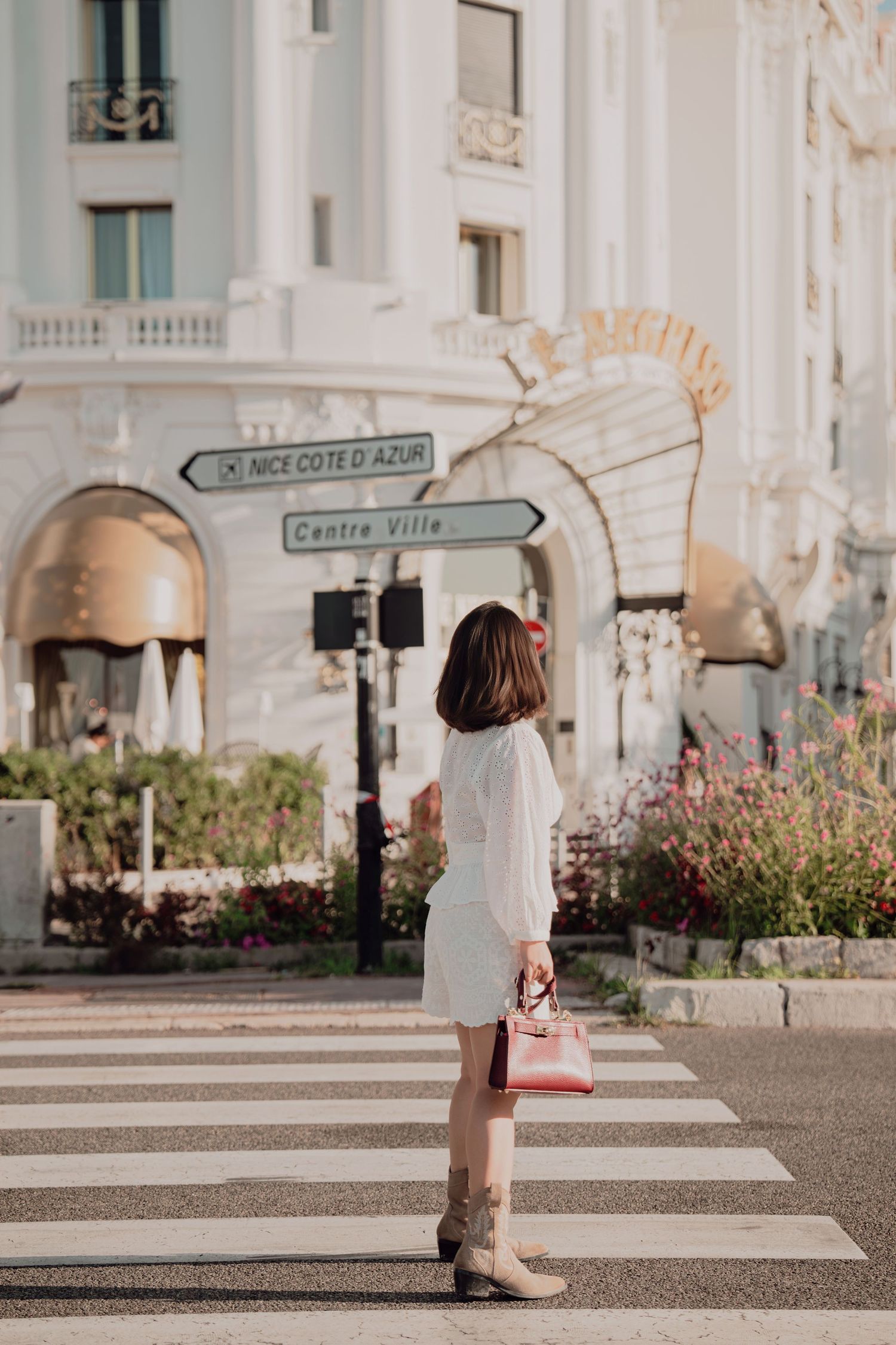 Woman in white dress with burgundy bag standing at crosswalk near Nice Cote d Azur sign.