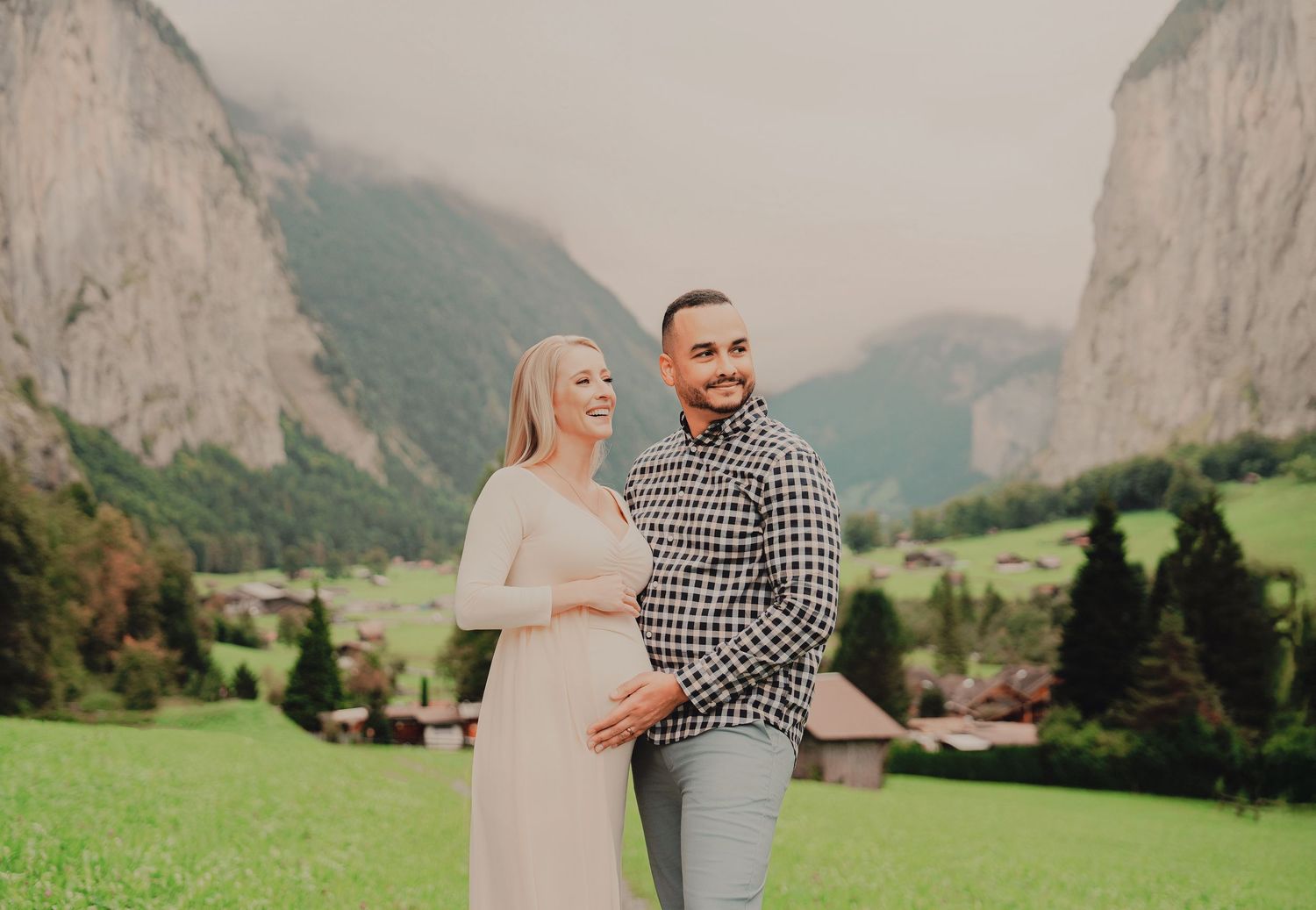 Expecting couple standing together in green Swiss alpine meadow with dramatic mountain cliffs in background.
