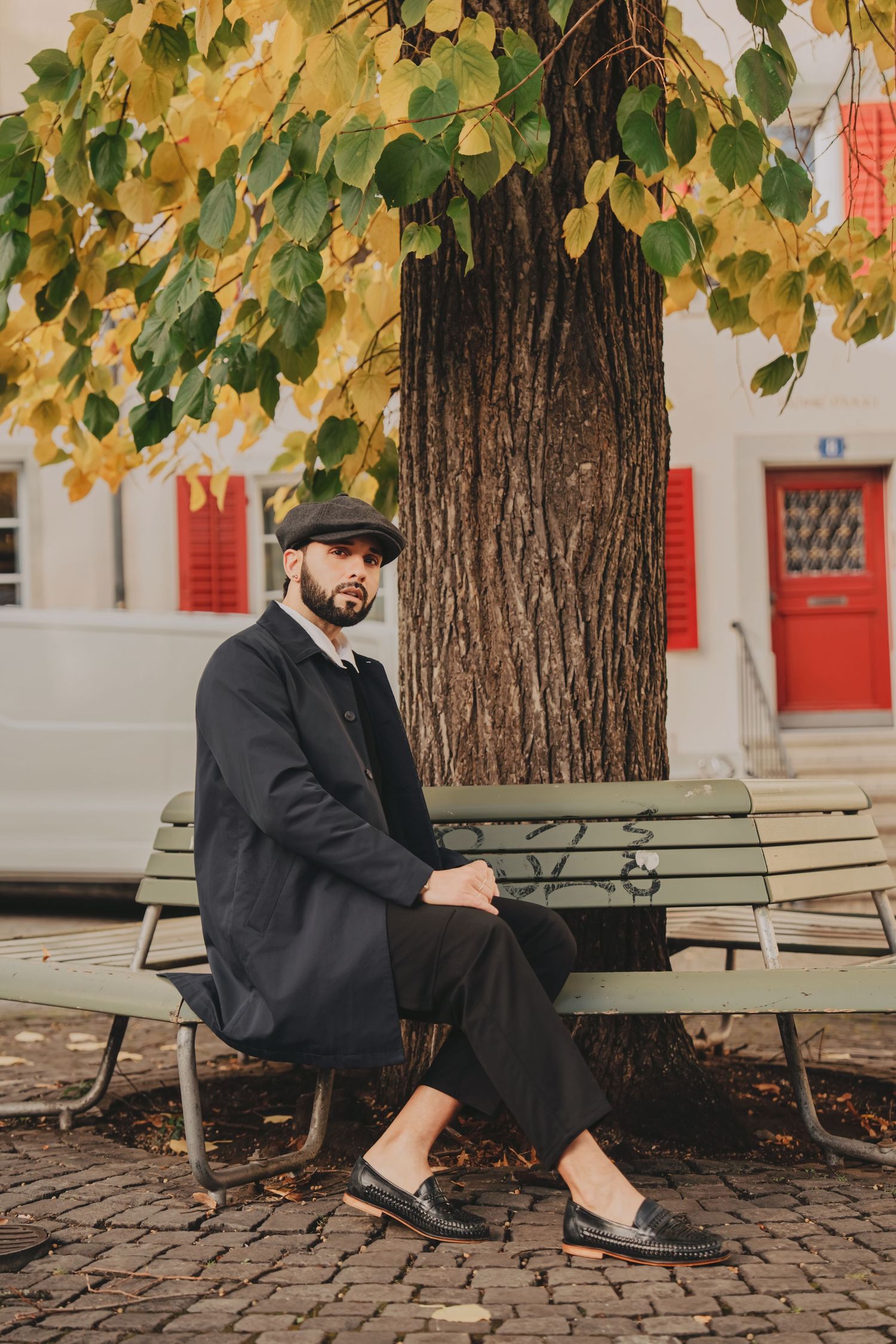 Bearded man in black coat and flat cap sitting on a bench under autumn trees in a European city square.