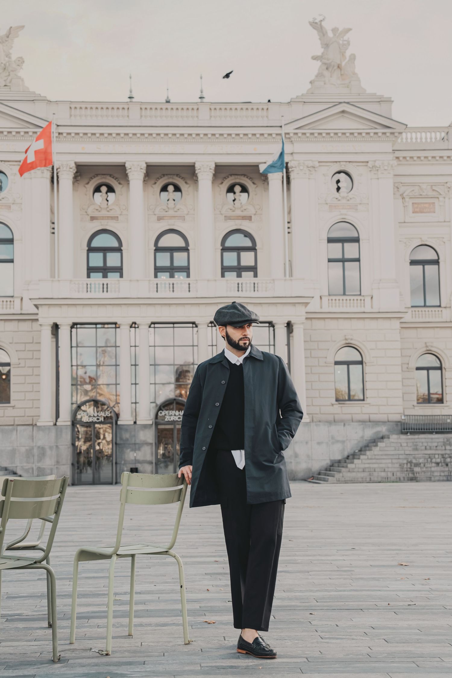 Fashionable man in dark coat and newsboy cap walking in plaza before grand neoclassical building.